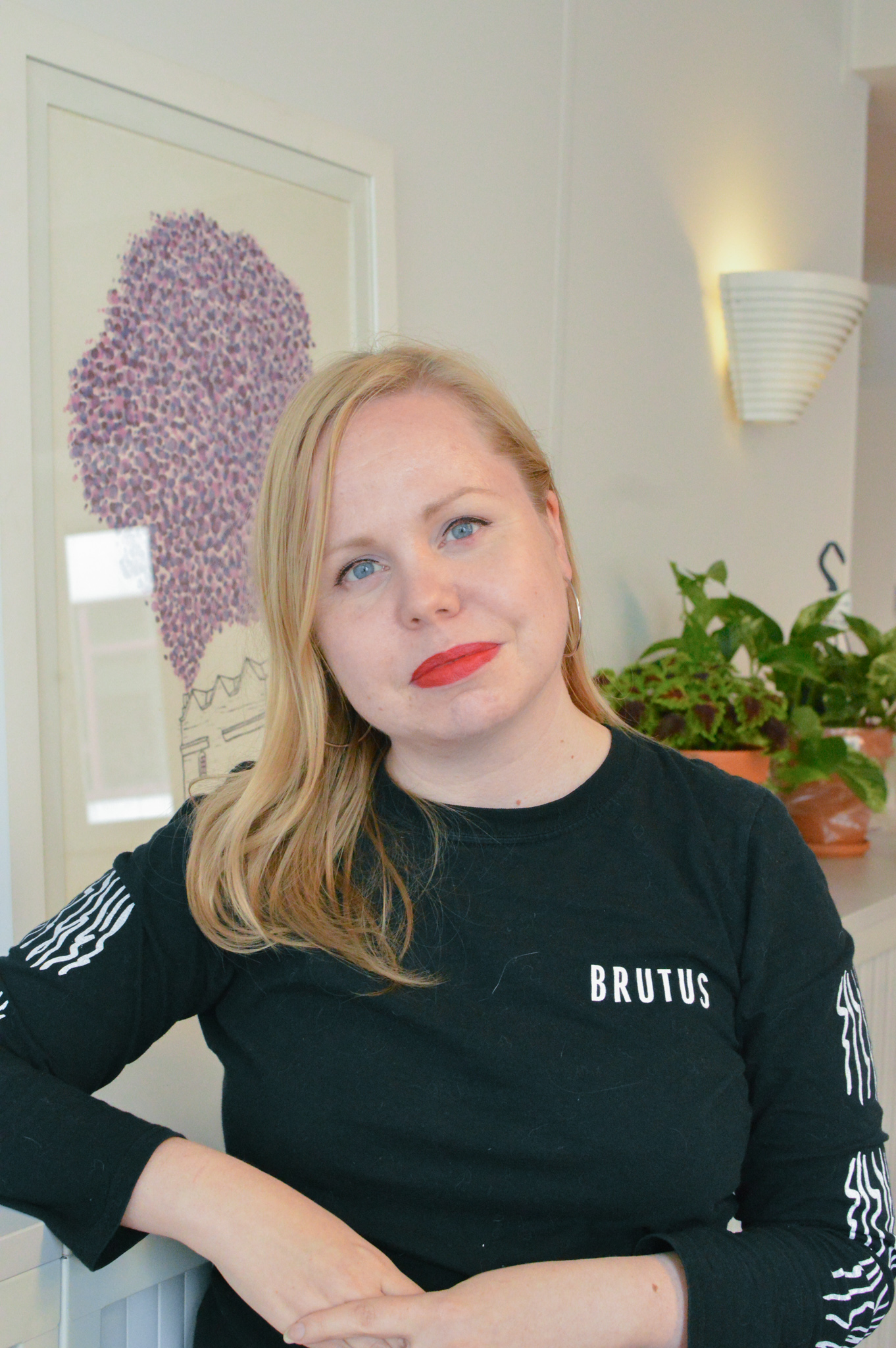 Malin Bergström_FCI_Agneskena Woman with long blonde hair, wears a black long sleeve top and leans against a cupboard.