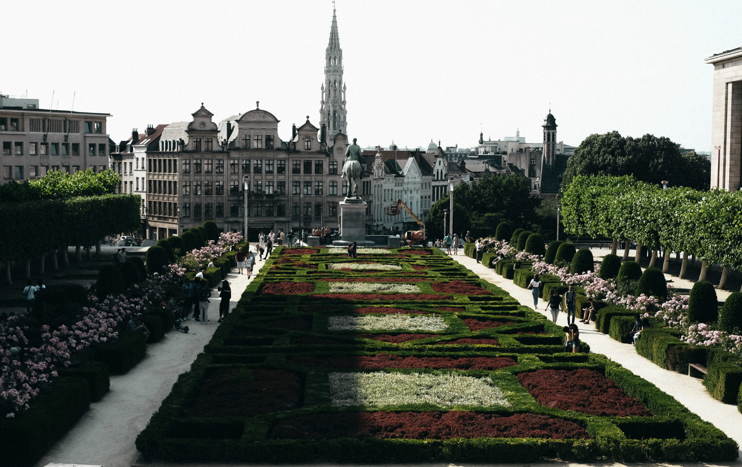 A symmetrical garden with neatly trimmed hedges and vibrant red, green, and white flower beds stretches toward a statue on a pedestal. The garden is flanked by pathways lined with blooming pink flowers and manicured shrubs, where people are strolling and sitting on benches. In the background, historic European buildings with ornate facades rise, including a towering Gothic-style spire. The scene is bathed in sunlight, casting long shadows across the landscape.