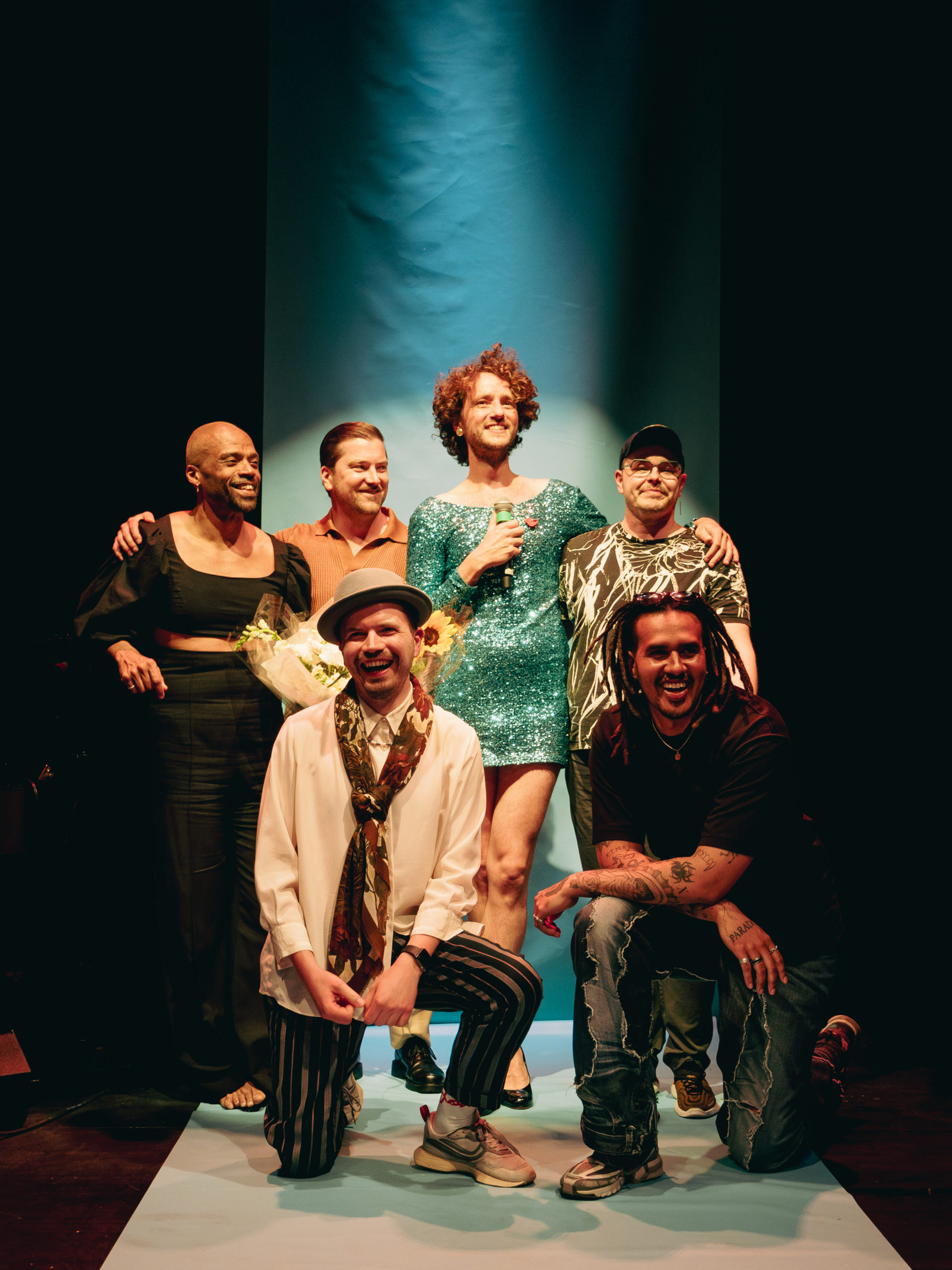 Cast members of Boys Won't Be Boys Finland edition come together while the audience members applaud at Svenska Teatern. The background includes a turqouise banner and theatre lighting.