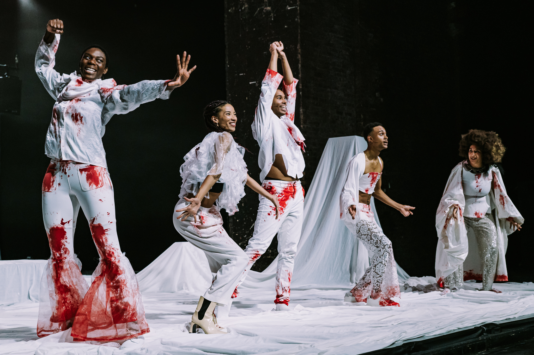 6 Black women and men stand on stage in a line in different stances of dance. They are all dressed in white and covered in red stains reminiscent of blood.