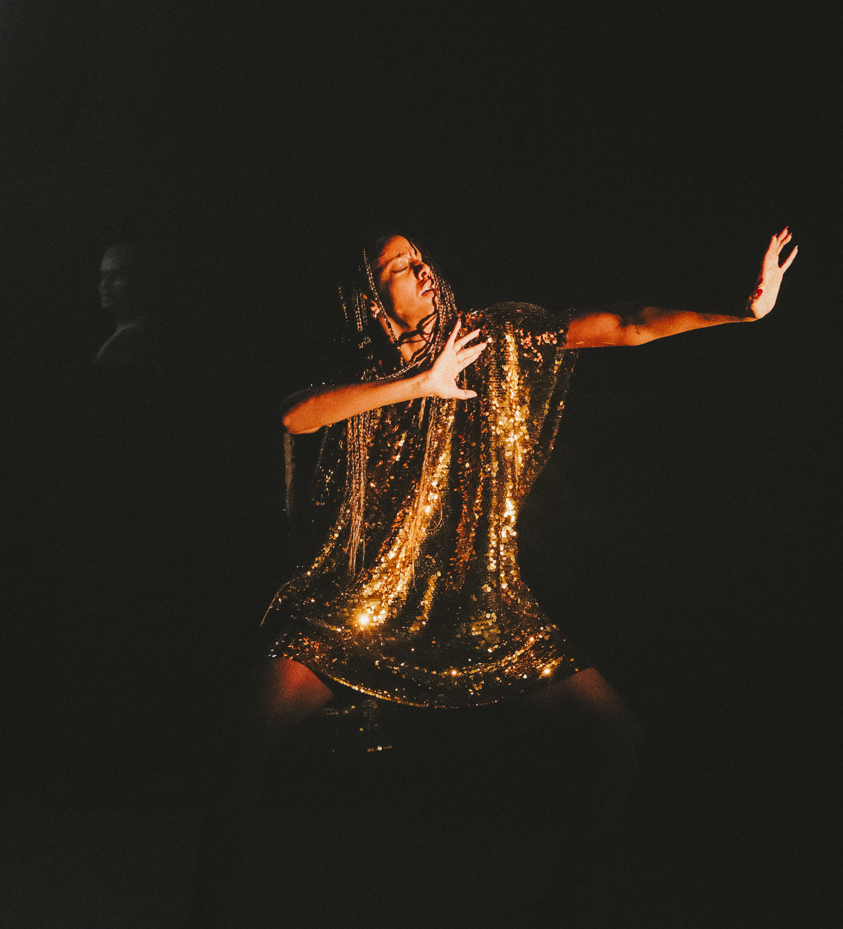 A black woman stands with her eyes closed, her arms are stretched out at chest hight with palms facing the right. She is dressed in gold. The light shines on her as the rest of the stage is in darkness. A figure can be seen behind her to the left in the shadows.