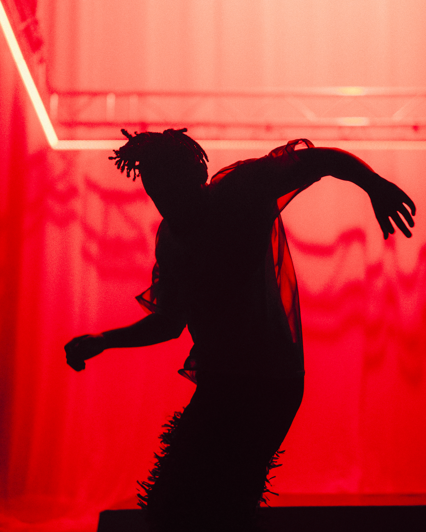 A black man stands with his arms stretched out to either side, making a wave-like pattern. The stage is strongly lit with a bright red tone.