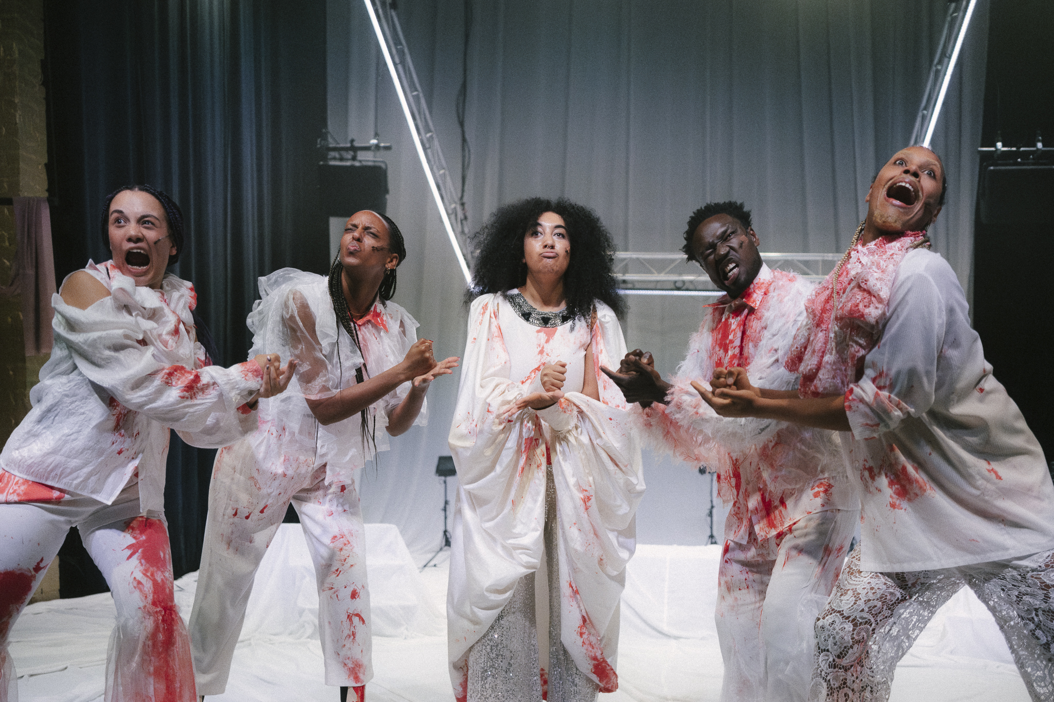 Five black performers play rock paper scissors on stage while making exaggerated facial expressions. They are dressed in white clothing with red, blood-like splatters on them.