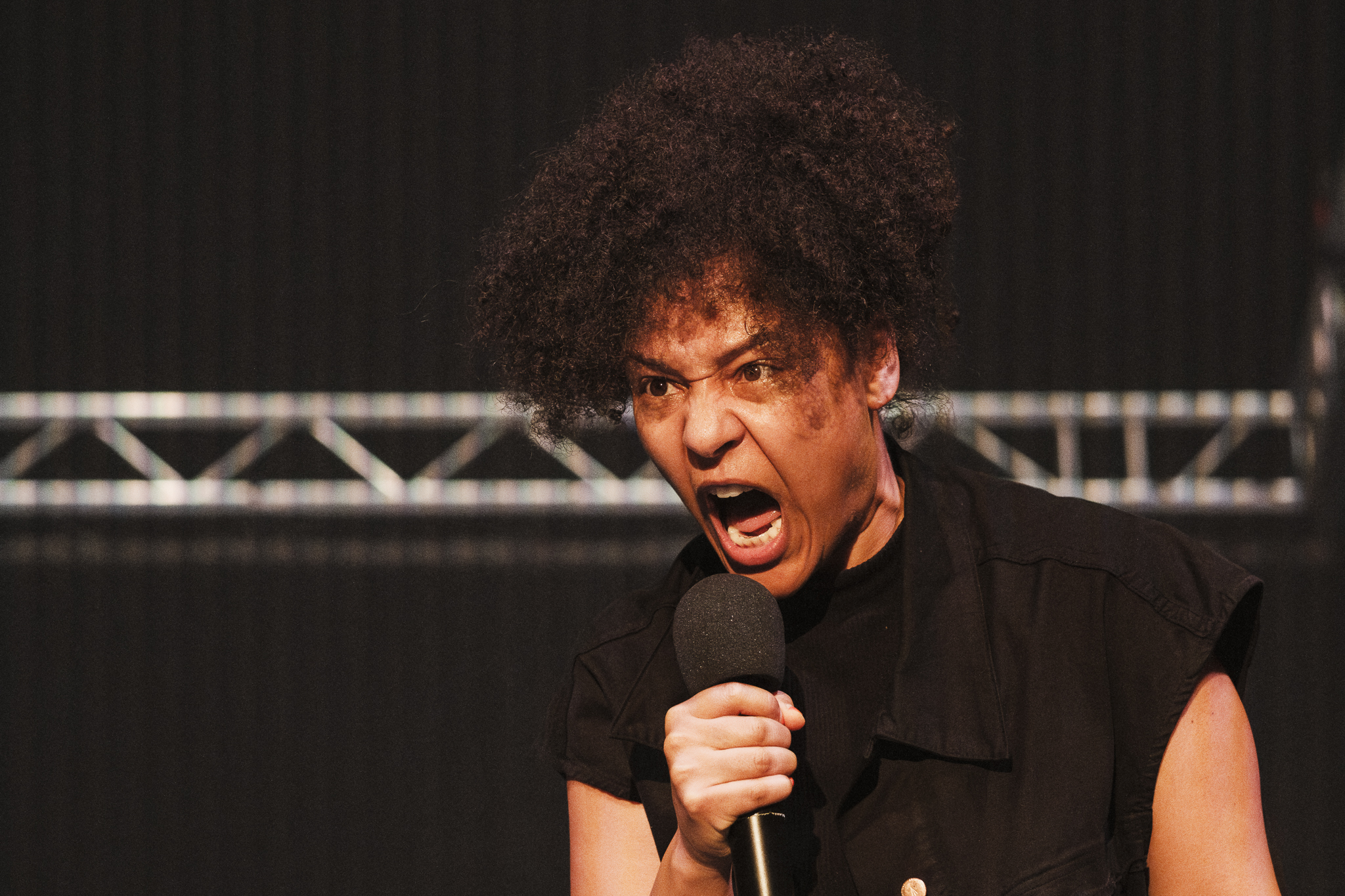 A black woman holds her mouth open while holding a microphone in front of her face. Behind her, a metal structure hangs against a black background.