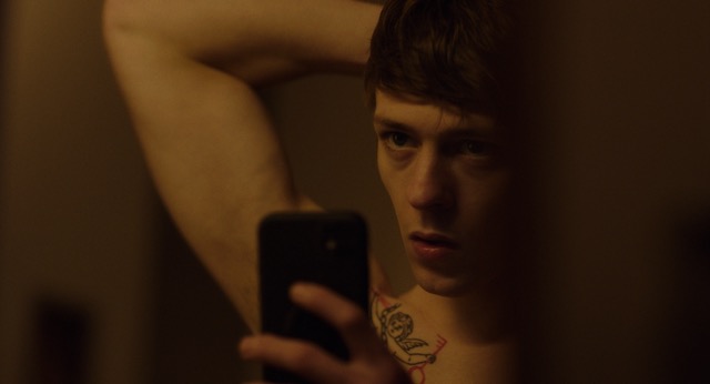 A man lies back topless. He has his right arm up resting behind his head. He holds his phone up in from of him, while glancing across the room.