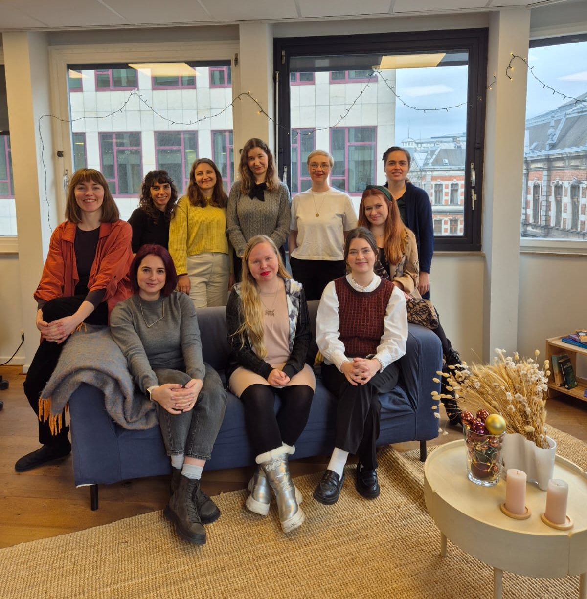 Ten women sit and stand around a blue couch in front of a window. They are smiling and looking straight at the camera.