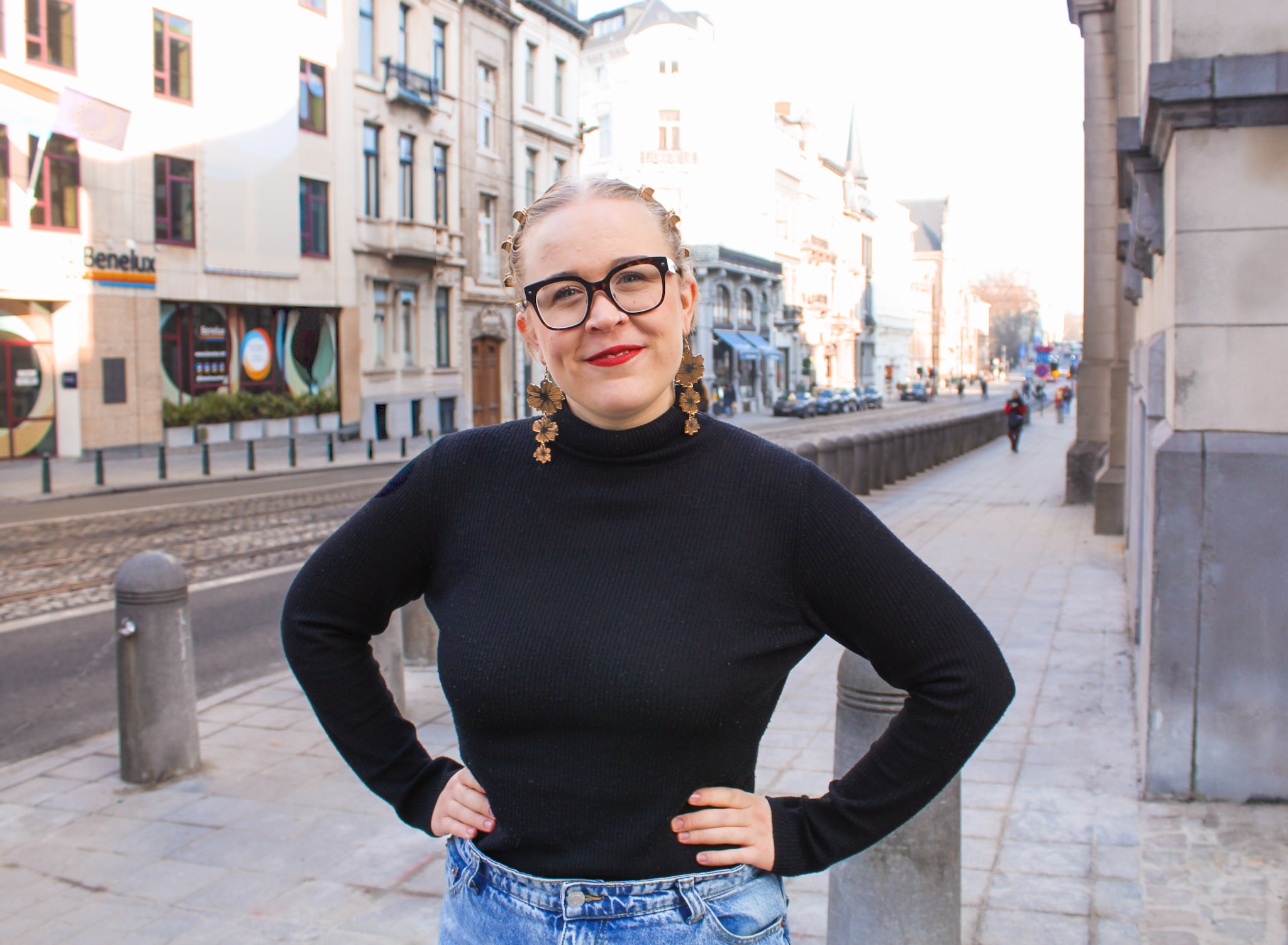 A white woman stands in the street with her hands on her hips and smiles at the camera. Her blond hair is clipped back. She wears glasses and dangly earrings. She wears a black turtleneck top and the top of her jeans can just be seen at the bottom of the photo.