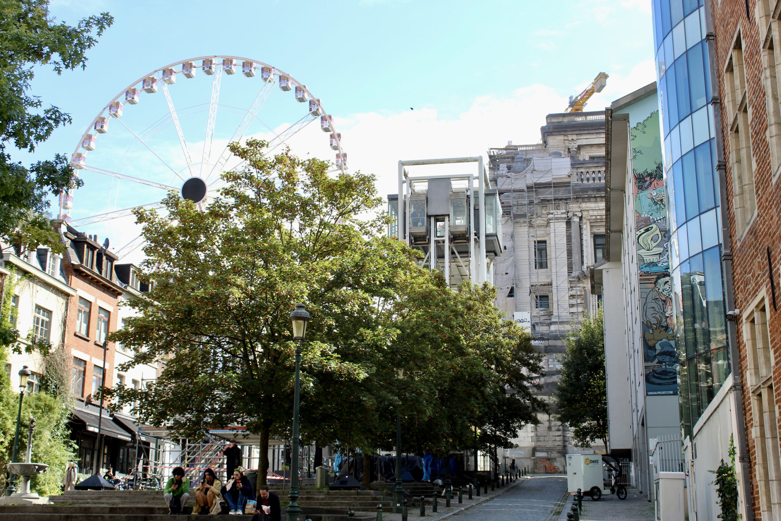 A city scene with a large Ferris wheel in the background, partially obscured by trees. In the foreground, there are several people sitting on steps under the shade of the trees. To the right, there is a modern glass building with a colorful mural on its side. Adjacent to it, an older, more classical building is under renovation, with scaffolding visible. A small street runs between the buildings, with a few bollards and a parked scooter. The sky is clear and blue, suggesting a sunny day.