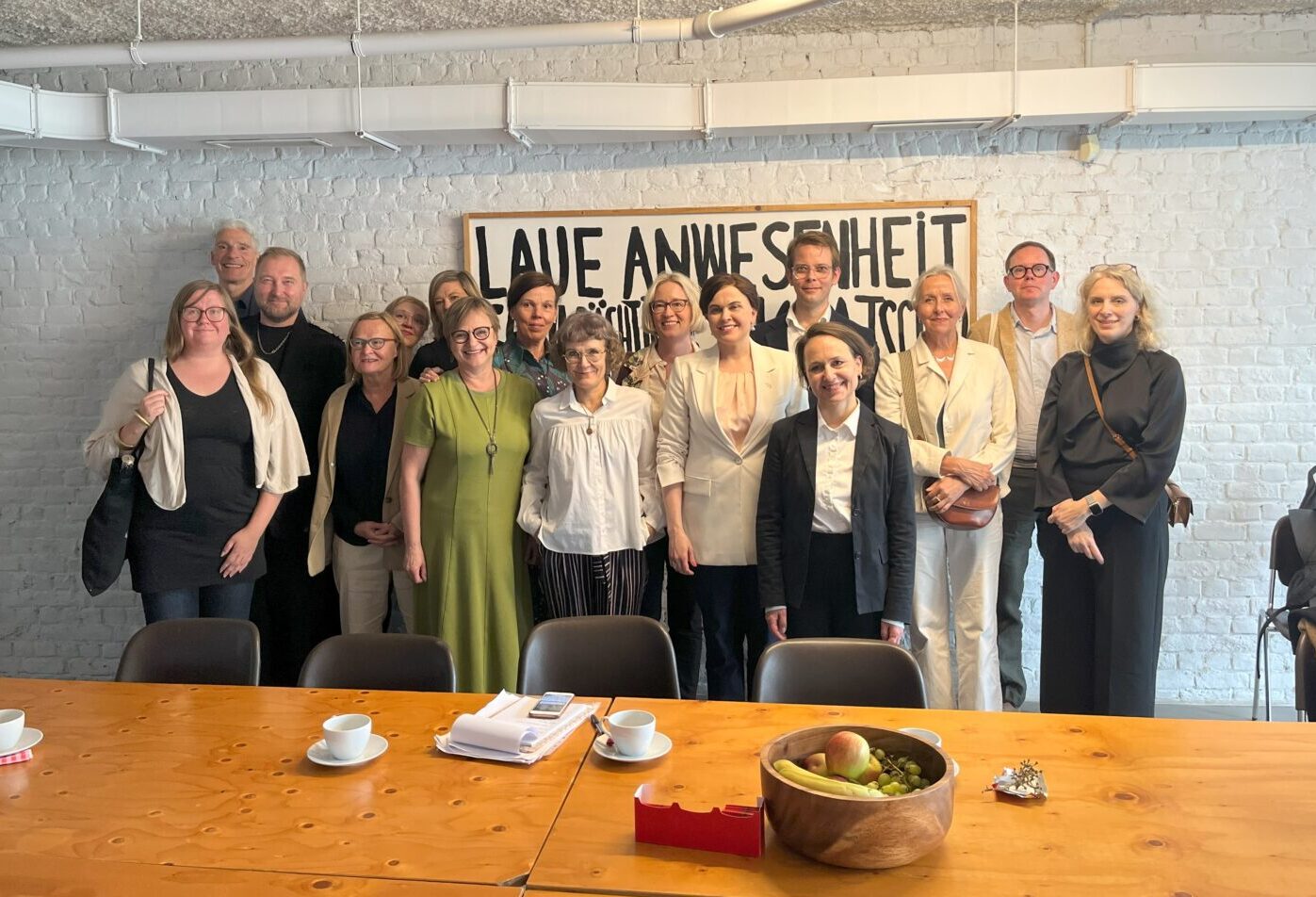 Group of people stand in front of a wall behind a large meeting table.