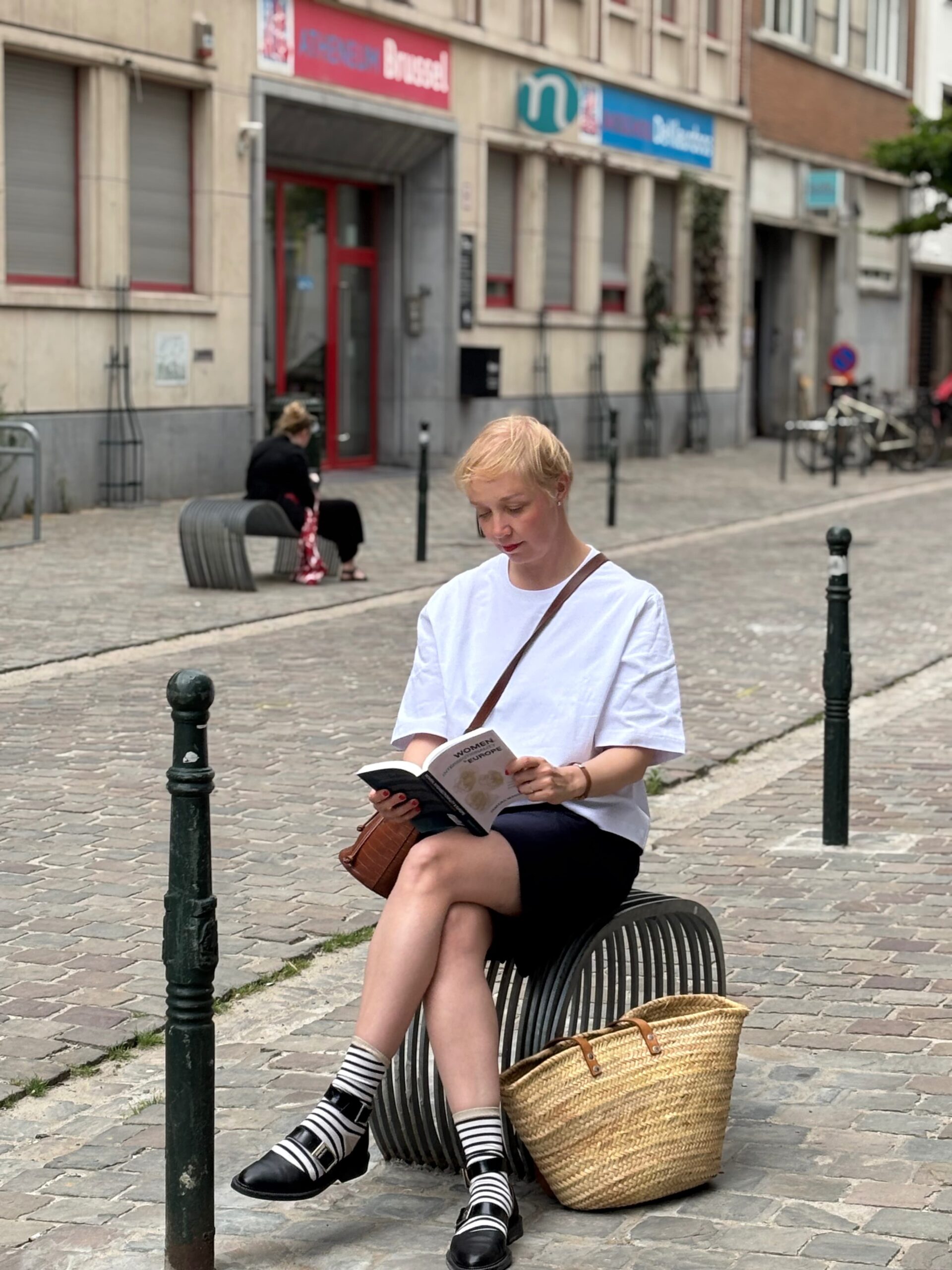 A woman sits on a metal bench in a quiet street. She is reading a book. She has short, blond hair and is wearing a white T-shirt, black shorts, stripy socks and cool black shoes. She also wears a brown leather cross body bag and has a basket bag down by her feet.