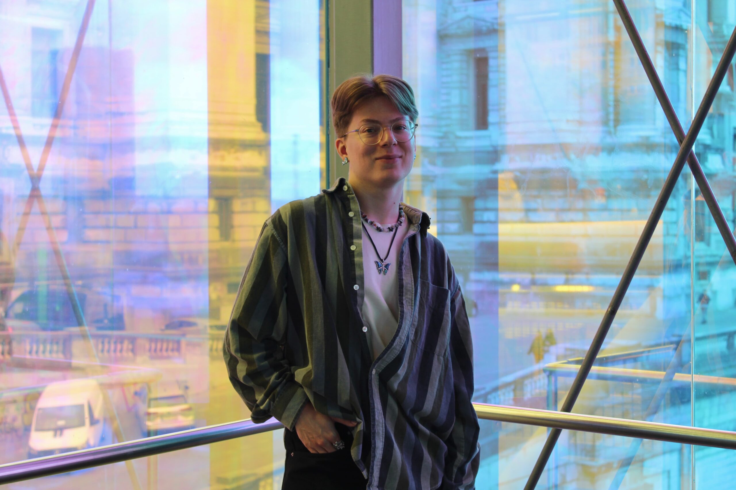 Oscar Arminen stands in a relaxed manner in loose striped clothing and layered jewelry, set against a backdrop of architectural structures seen through shimmering blue-yellow glass.
