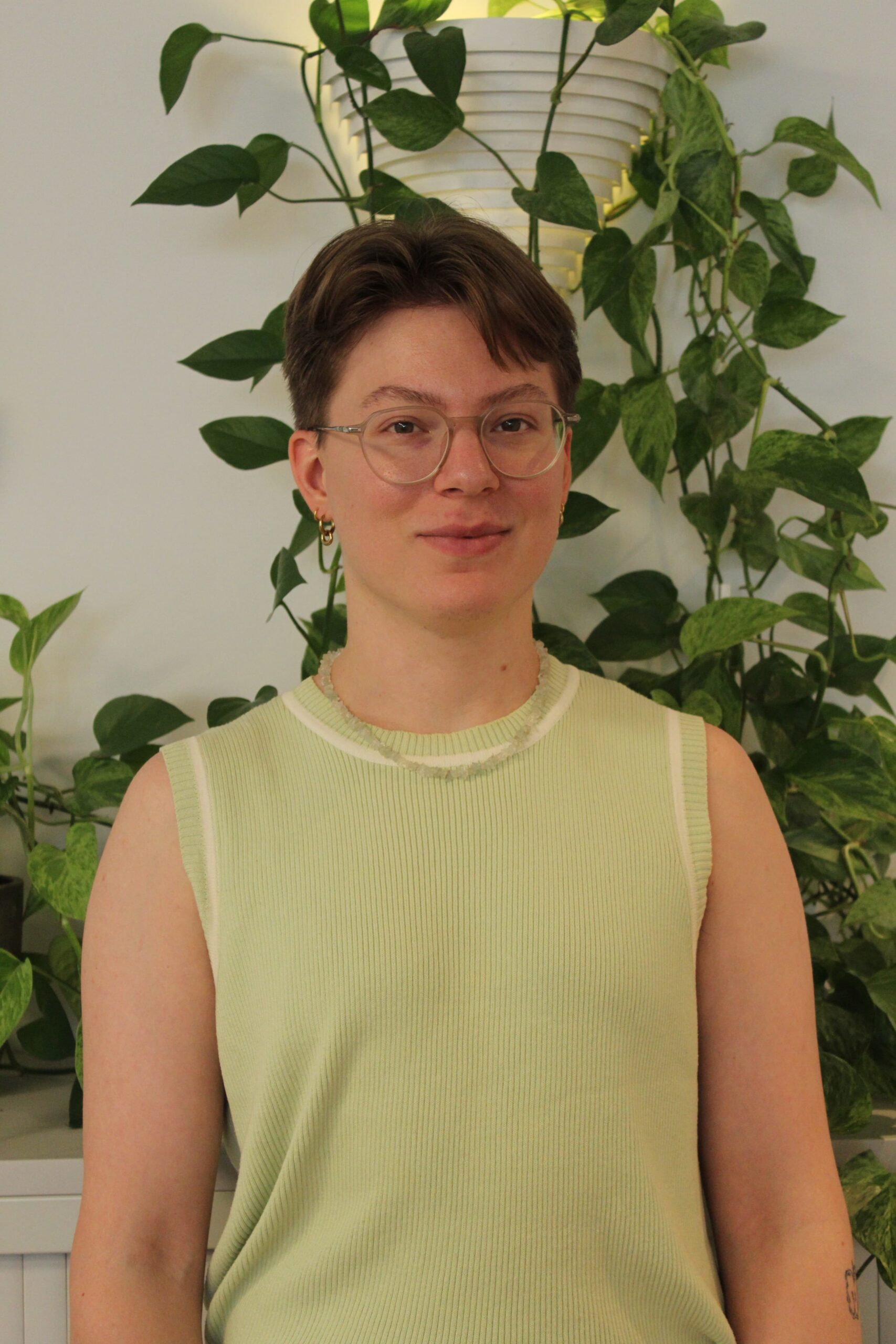 Oscar Arminen with short brown hair and glasses smiles gently, wearing a sleeveless, knitted, light green top and standing in front of a wall covered with leafy green vines.