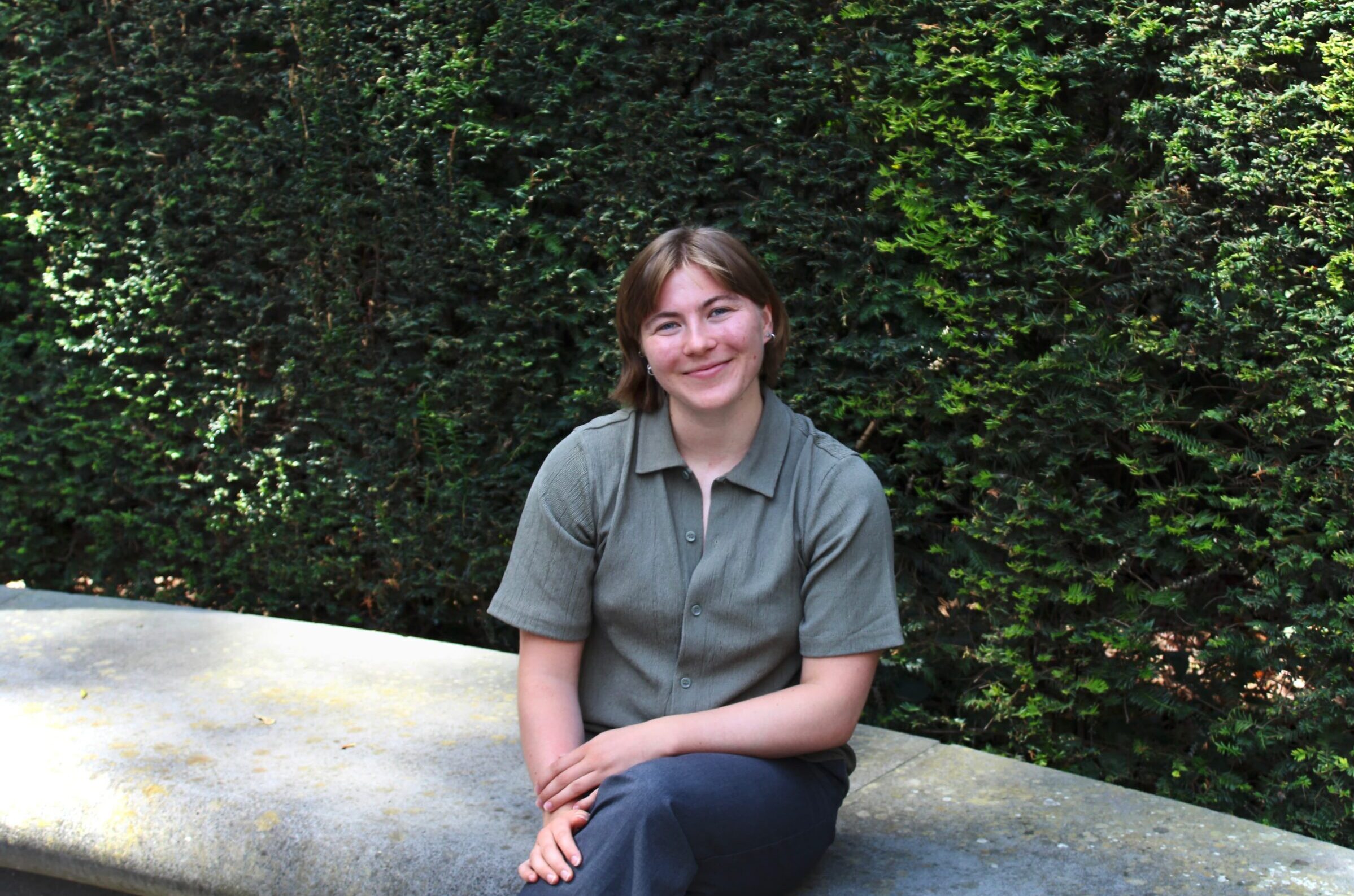 Tilda Forss, sits cross-legged on a stone bench, smiling at the camera. There is a tall green hedge in the background. They have shoulder length, light brown hair and are wearing a light green short-sleeved button-up shirt and dark trousers.