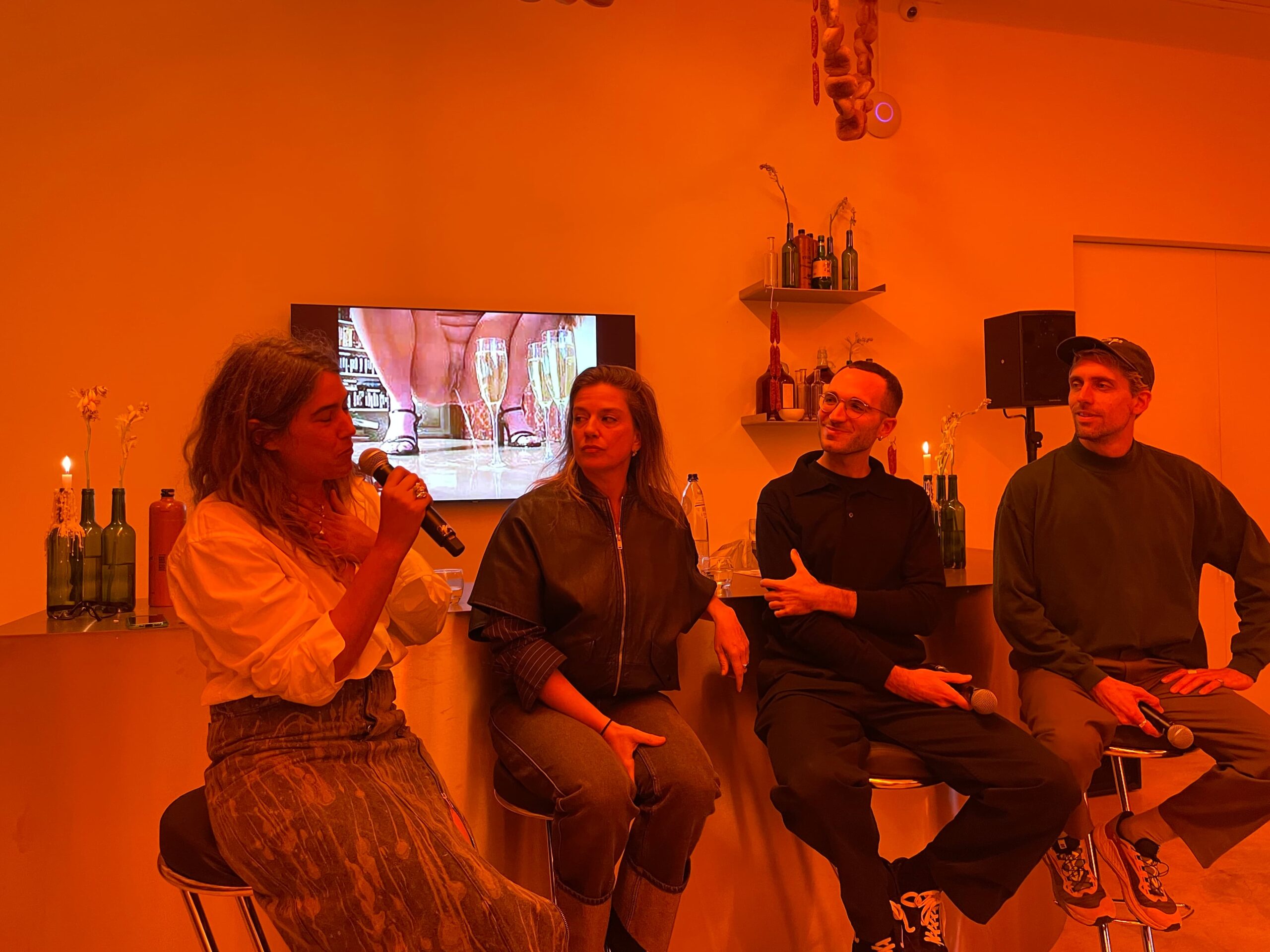 An intimate panel discussion with four speakers seated side by side, warmly lit in orange, with a screen showing an art video and bottles with candles placed nearby.
