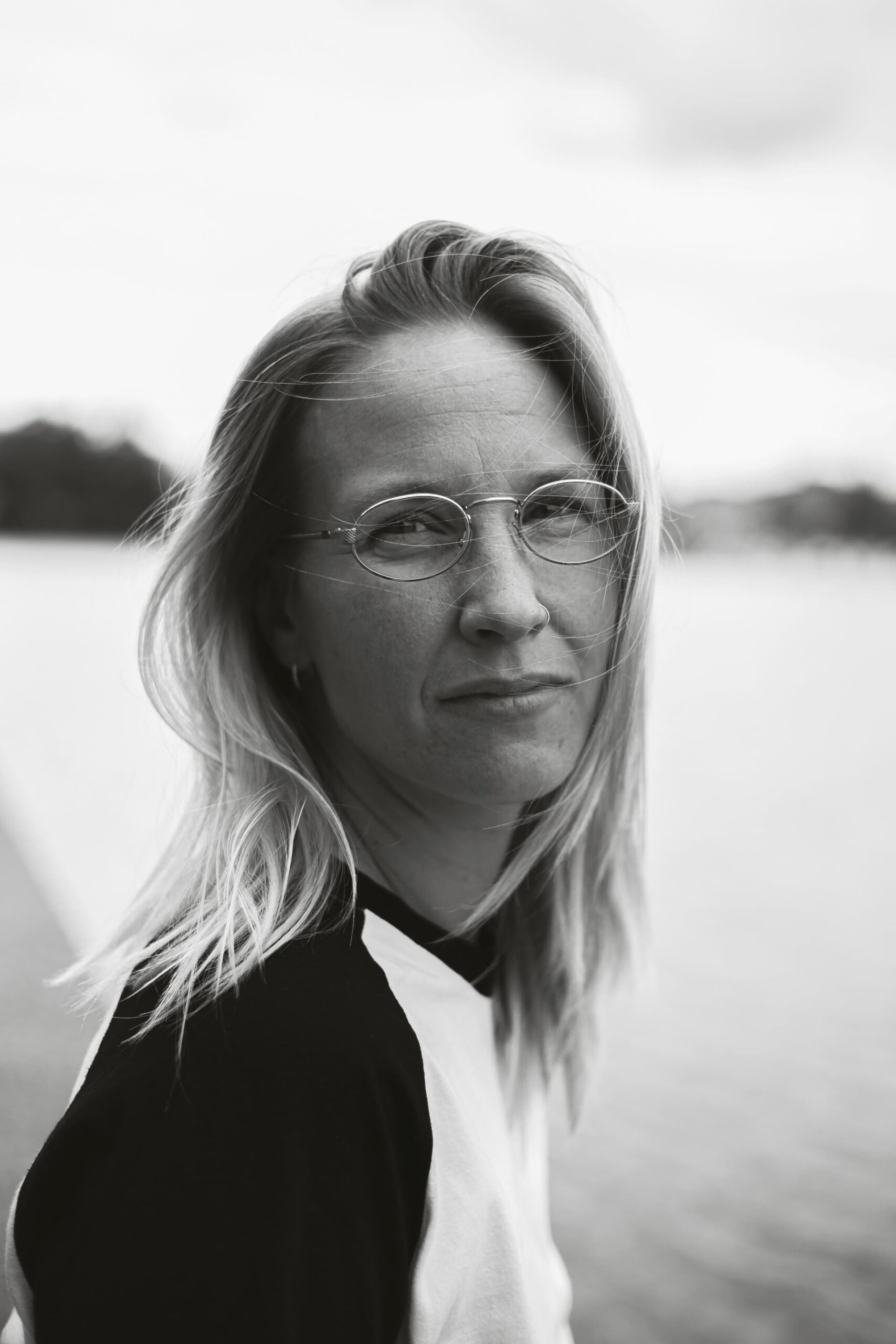 Max Hands looks directly at the camera. They have shoulder length hair and are wearing glasses and have a nose ring. The portrait is in black and white. 