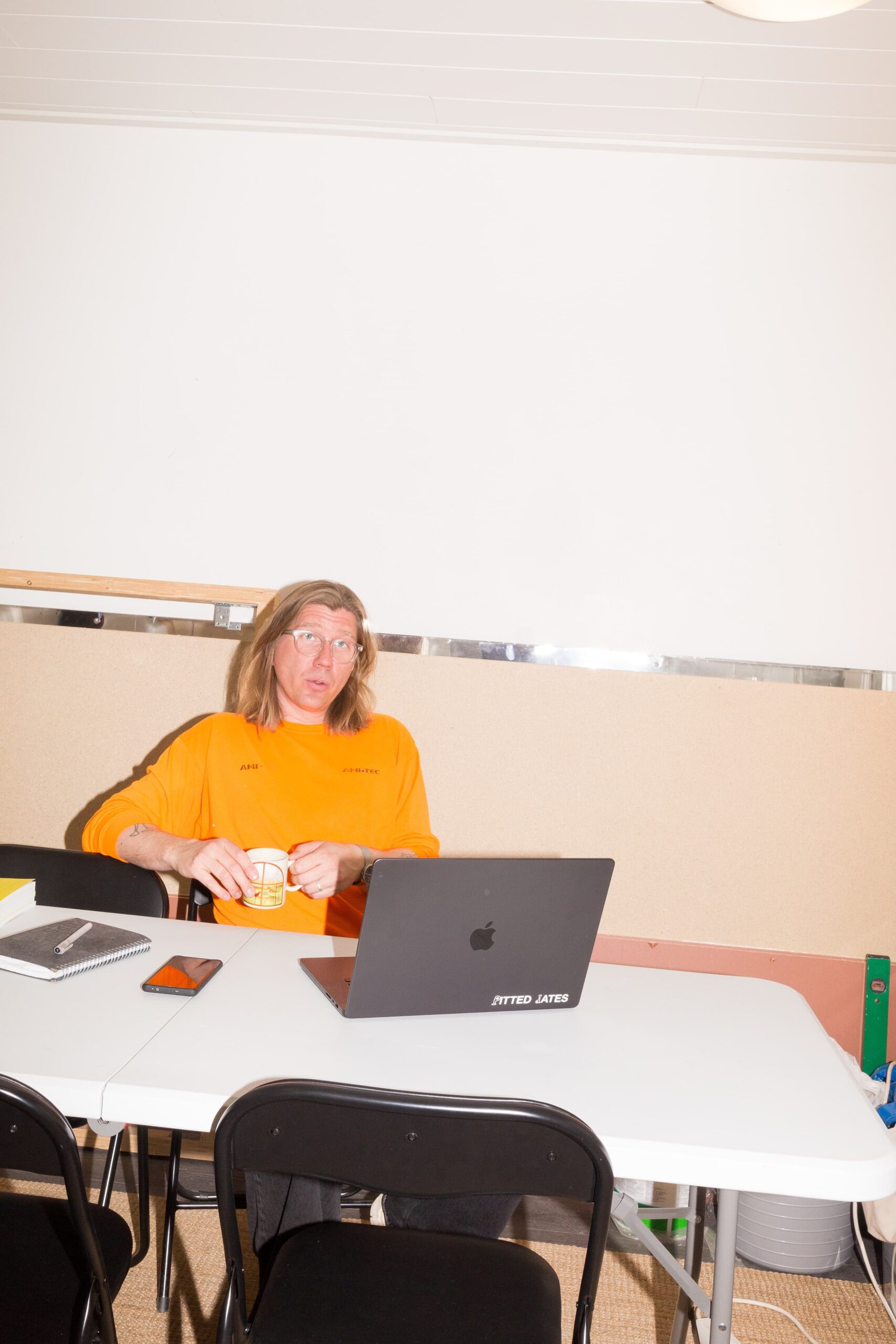 A man with shoulder length light coloured hair sits at a table with a laptop. 