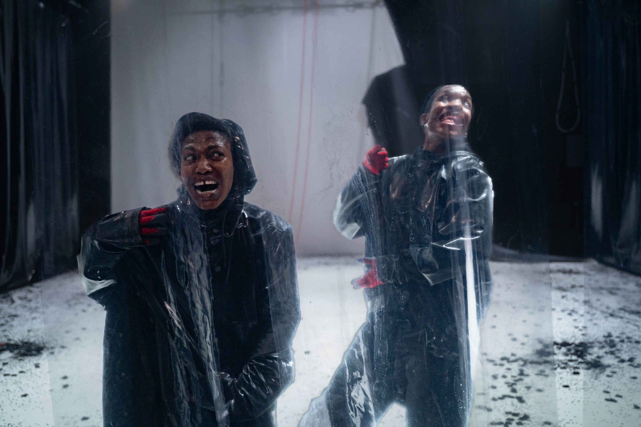 Two Black people wearing black raincoats and red gloves stand behind semi-transparent plastic sheets in a dimly lit space. One appears distressed, mouth open in an anguished expression, while the other looks joyful, mid-laugh. The floor is white with scattered debris, and dark curtains frame the background, creating a dramatic, theatrical atmosphere.