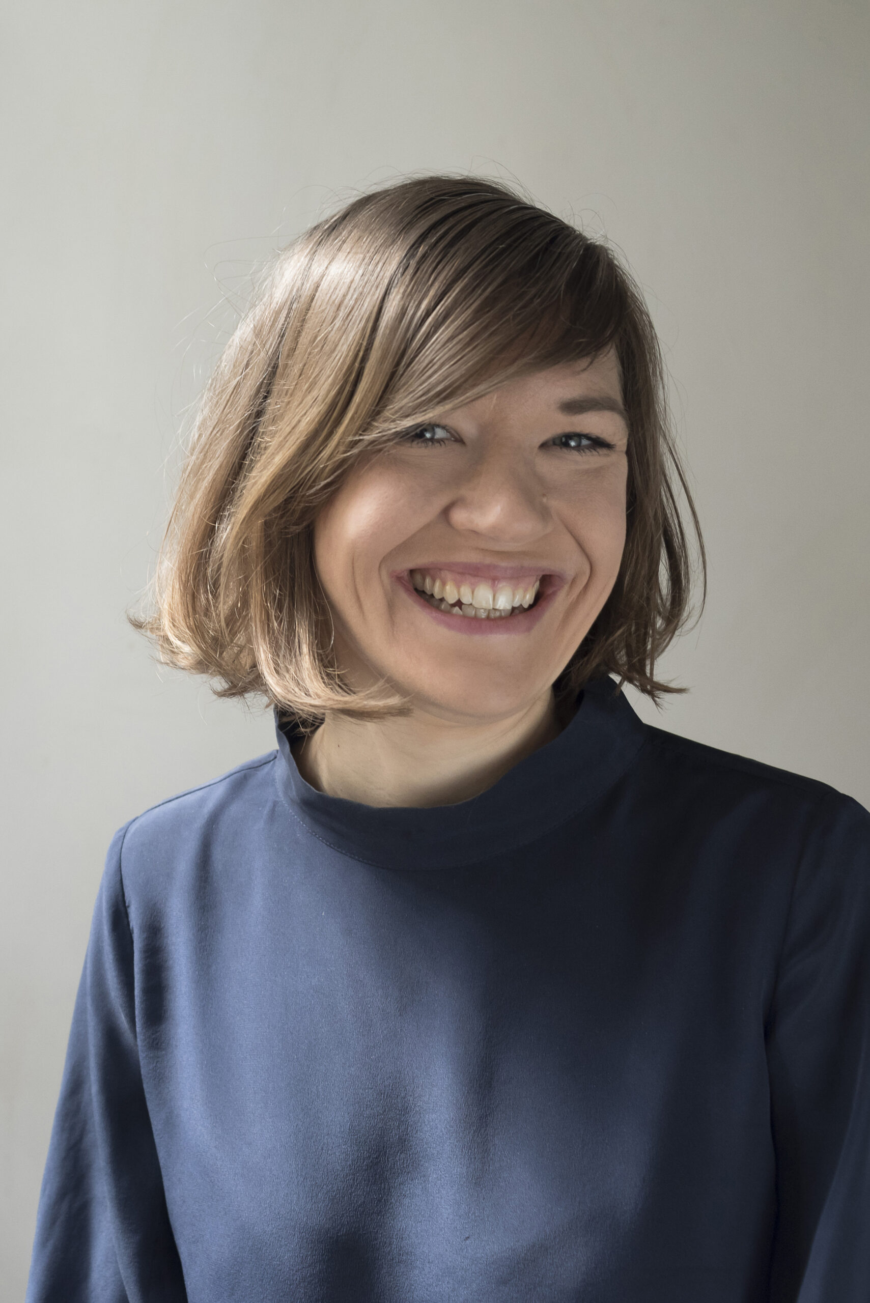 Johanna Råman ©_Alessandra_Carosi Portrait of Johanna Raman. Close-up of a smiling woman with side-parted brown hair and a dark blue outfit, photographed in front of a neutral wall.