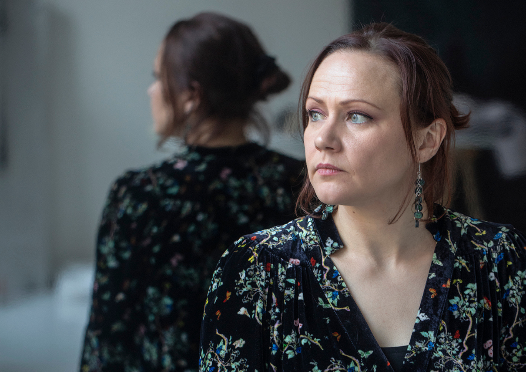Pyykkinen_Tiina_01-Photo-Jorma-Puranen-finncult-website Tiina Pyykkinen sits facing the camera but looking away to the side. There is a mirror behind her. She is wearing a black dress with a small flower print and long earrings with beads. She has light skin, brown hair up in a bun, and blue eyes.
