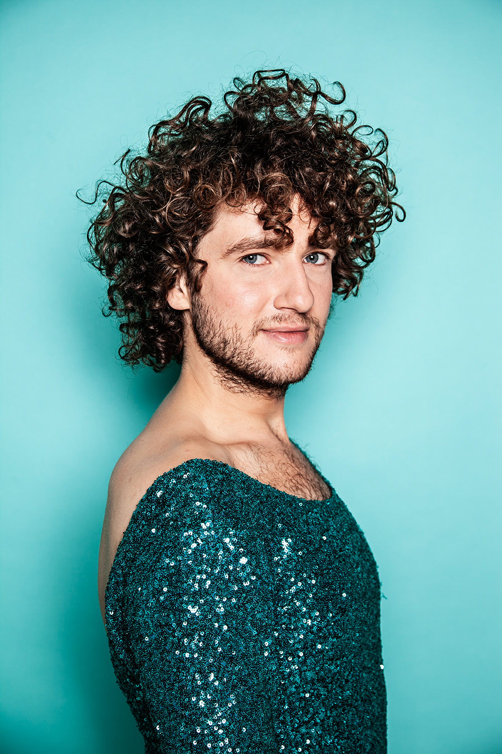 Rikkert van Huisstede looks into the camera in front of a teal colores backdrop. Rikkert has light skin, curly brown hair, a beard and blue eyes. He is wearing a light blue sequin dress.