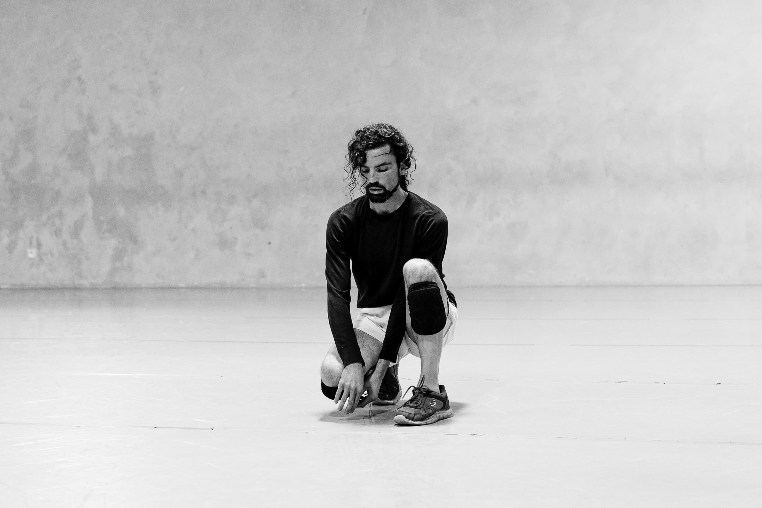 23_PER_Crude_Nov18_Pic (@ArnaudBeelen) A black and white photo showing Kevin Fay kneeling with one knee bent on a dance studio floor. He wears a long black top and white shorts. He has black knee pads on. His hair covers parts of his face. He has light skin, curly brown hair and a beard.