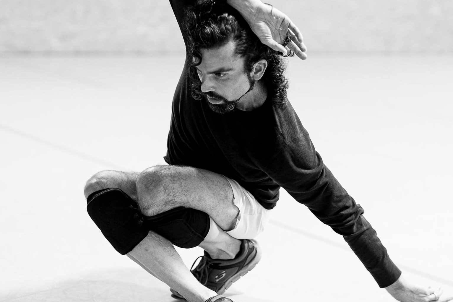 A black and white photo of a man performing. He crouches to the ground with one arm resting over his head. He wears a long black top and white shorts. He has black knee pads on.