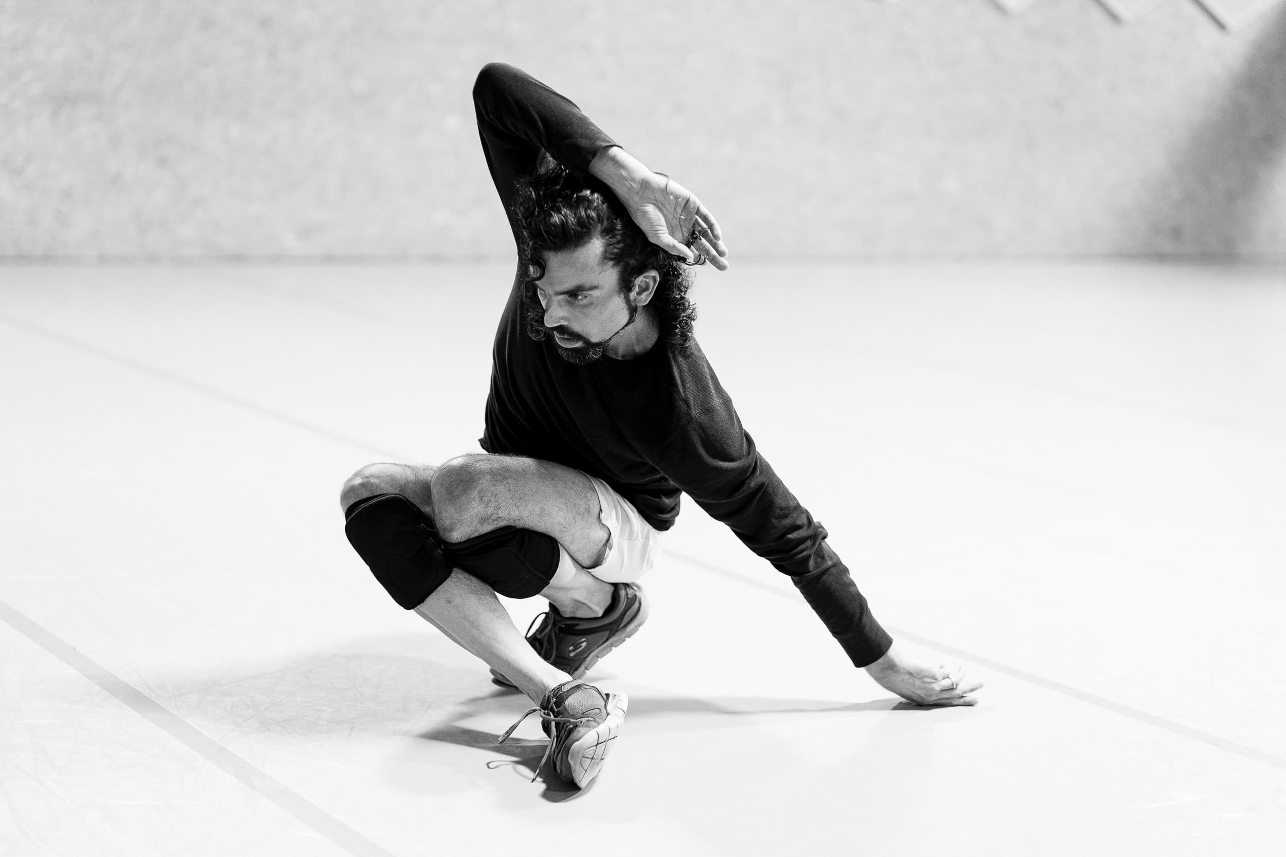 A black and white photo of a man performing. He crouches to the ground with one arm resting over his head. He wears a long black top and white shorts. He has black knee pads on.