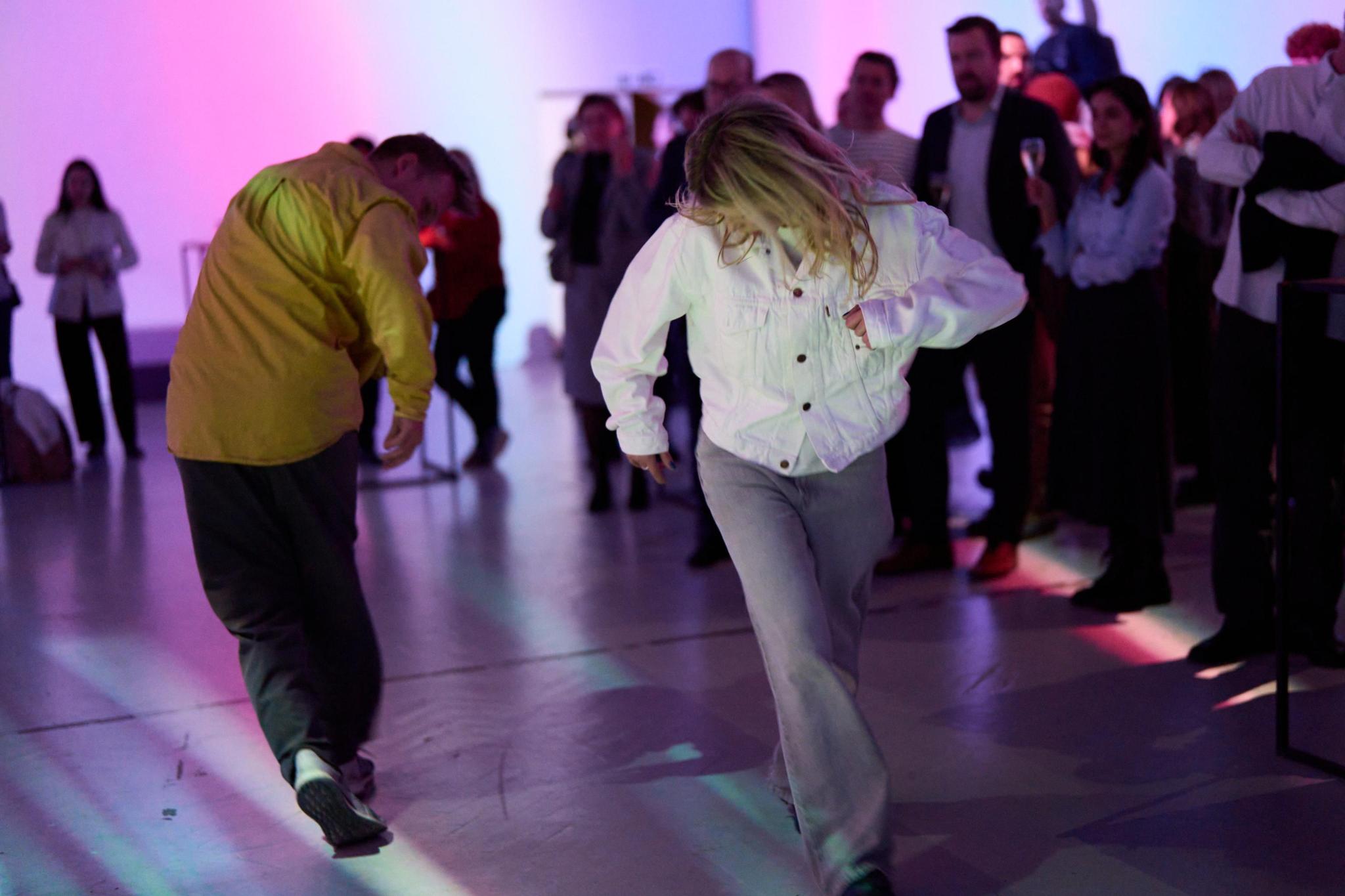 Two people are dancing. One is wearing a white denim jacket and grey jeans, and the other wears a yellow button-up and black pants. People are watching the performance from the side.