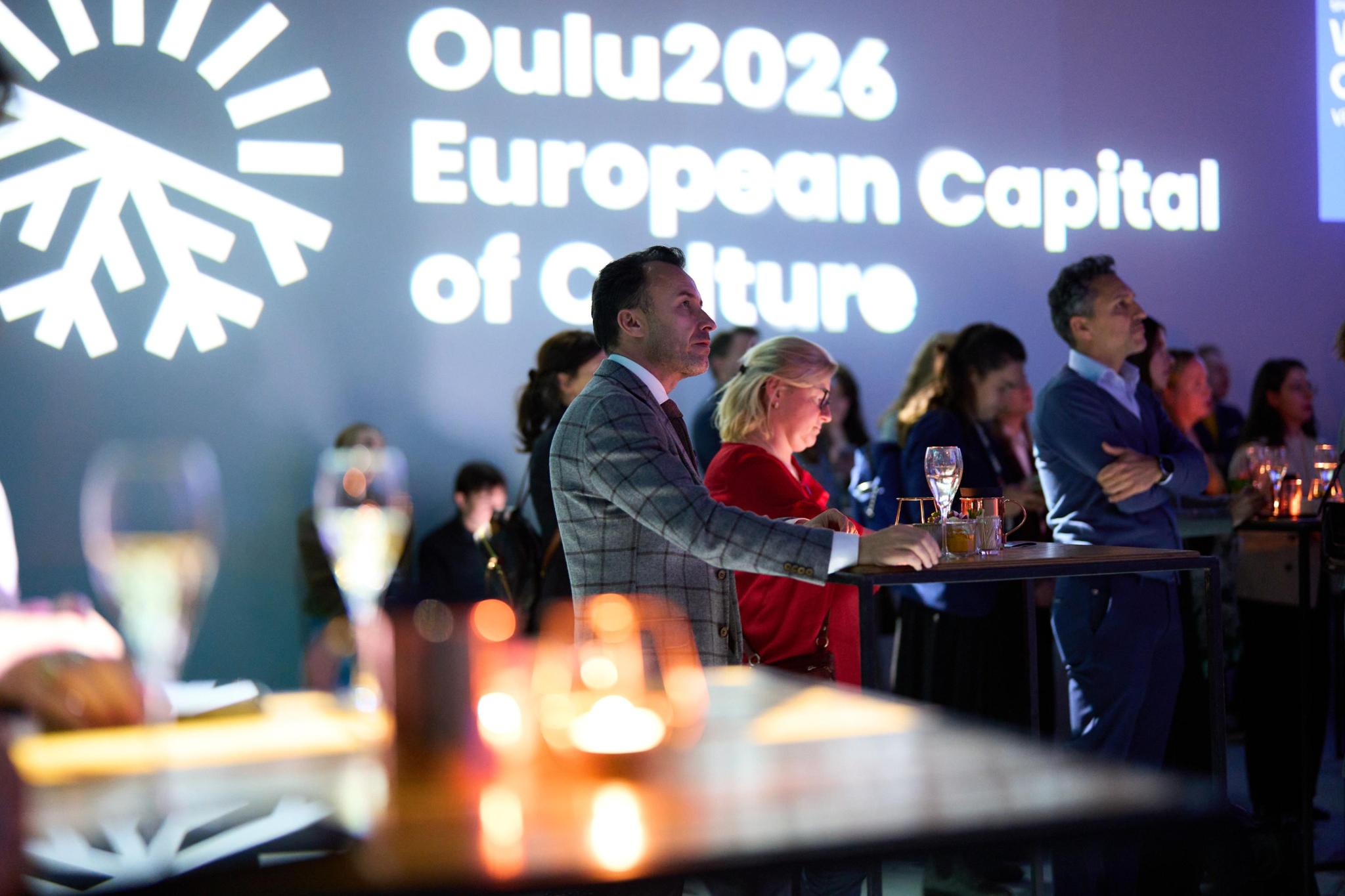 People stand in a darkened room with the text "Oulu2026 European Capital of Culture" projected on the wall.