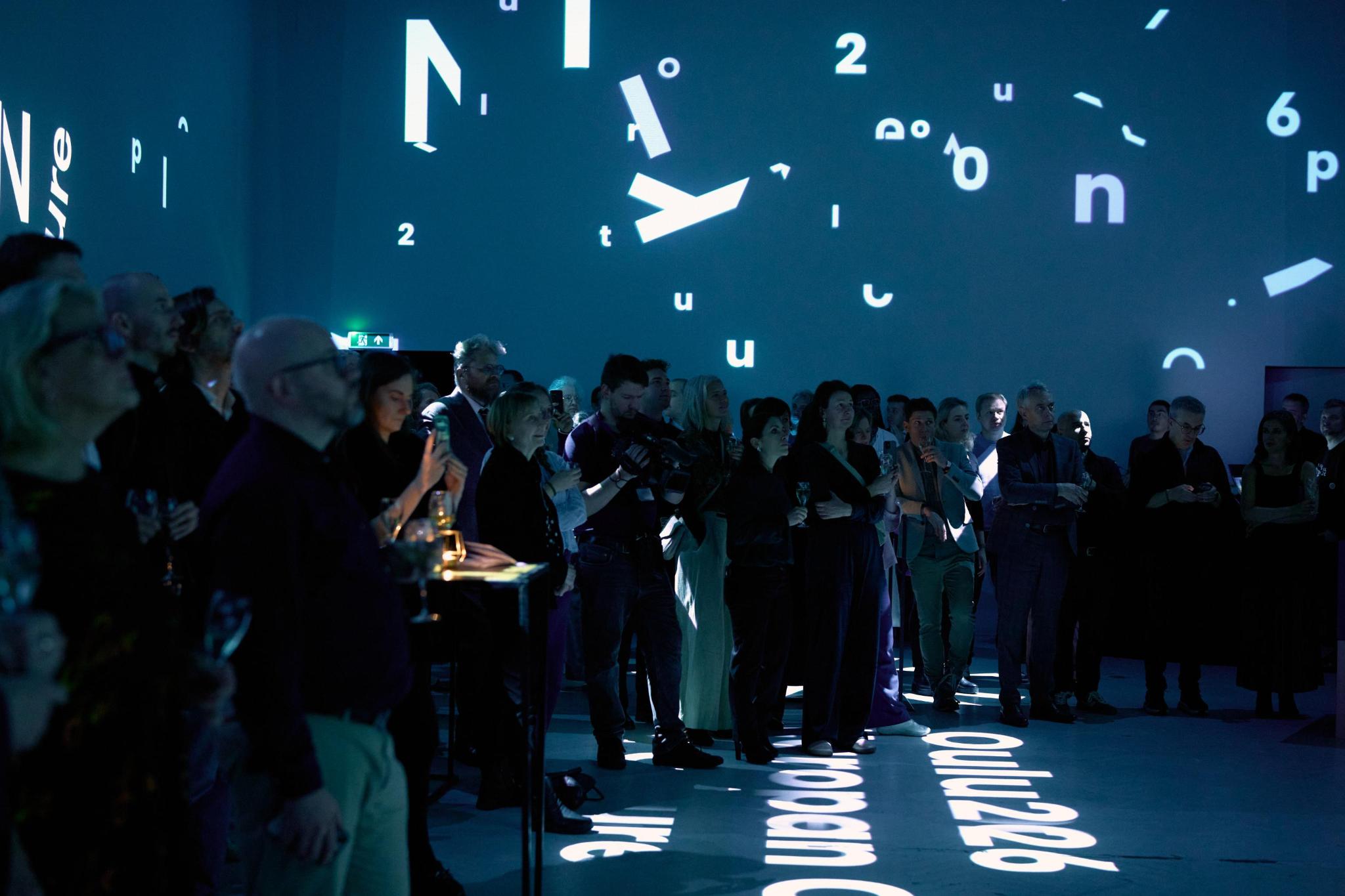 Oulu2026-photoby-Antonette de Groot People stand in a dark room with a video installation projected on the walls and floor. The floor reads "Oulu2026"