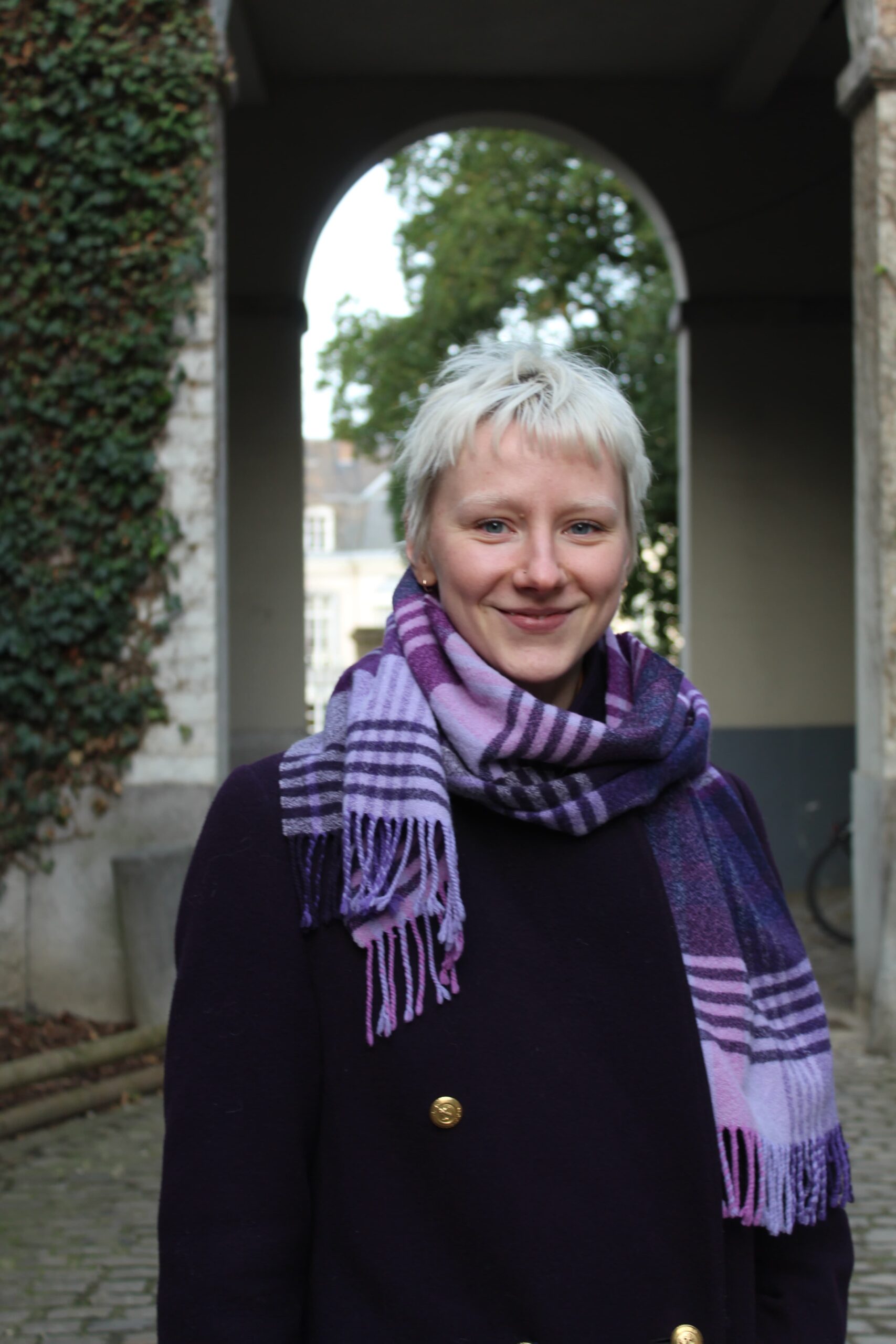 Margareeta stands smiling in a park with a white stone archway behind her. She has short blond hair and is wearing a dark coat and a purple plaid scarf.