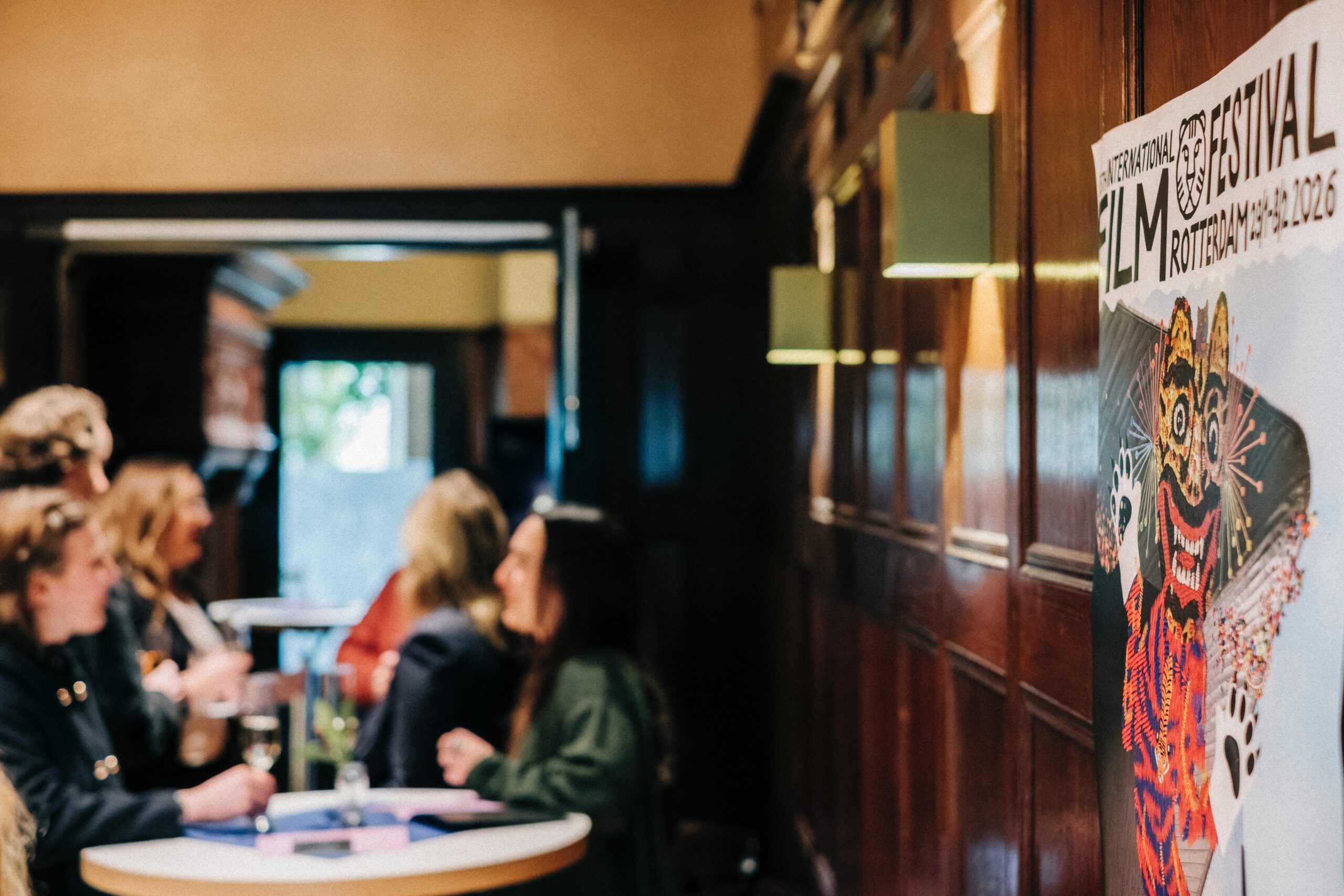 FCIxIFFR_2026_YohannesHenriksson-web A group of people are seen to the left, slightly out of focus. They are standing at high tables, chatting, with glasses of sparkling wine. To the right is a roll up poster for the International Film Festival Rotterdam.