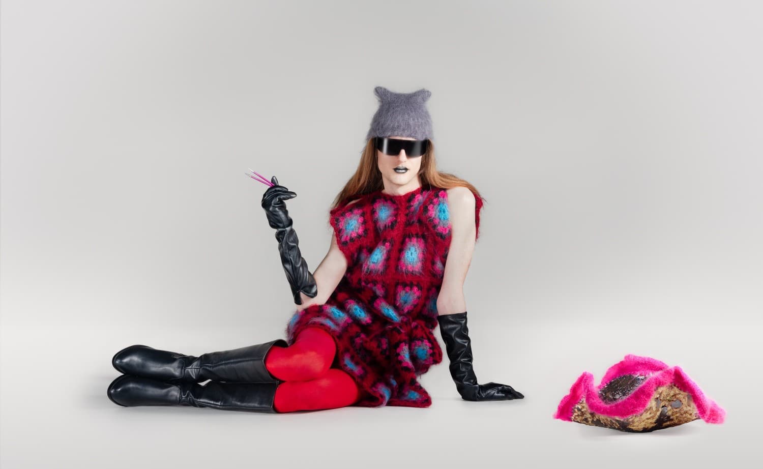Juha Vehmaanperä_ Interexterior Decoration 2026-Photo-Diana Luganski Juha-Vehmaanpera sits on the floor of a blank room. They are wearing a brightly coloured knitted dress, red tights, long black boots, a grey knitted hat, dark sunglasses and long black leather-like gloves. They have metallic lipstick on and are holding what appears to be two pink needles like a cigarette. There is a knitted item on the floor to the front right of them.