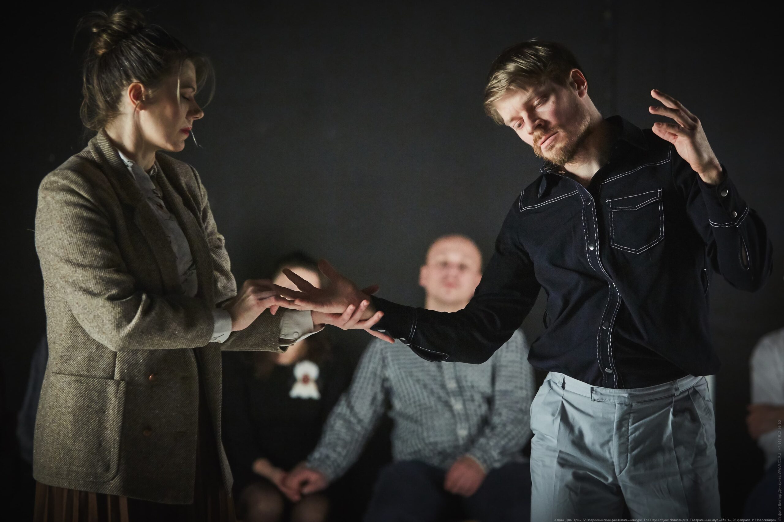 The Days Project. Maria Nurmela-Ville Oinonen. Photo by Victor Dmtriev A woman in a blazer stands to the left, eyes closed and gently strokes the hand of a man standing to the right. He reaches his hand out to her in movement. Behind is a dark wall and two members of the audience can be see sitting watching, holding hands.