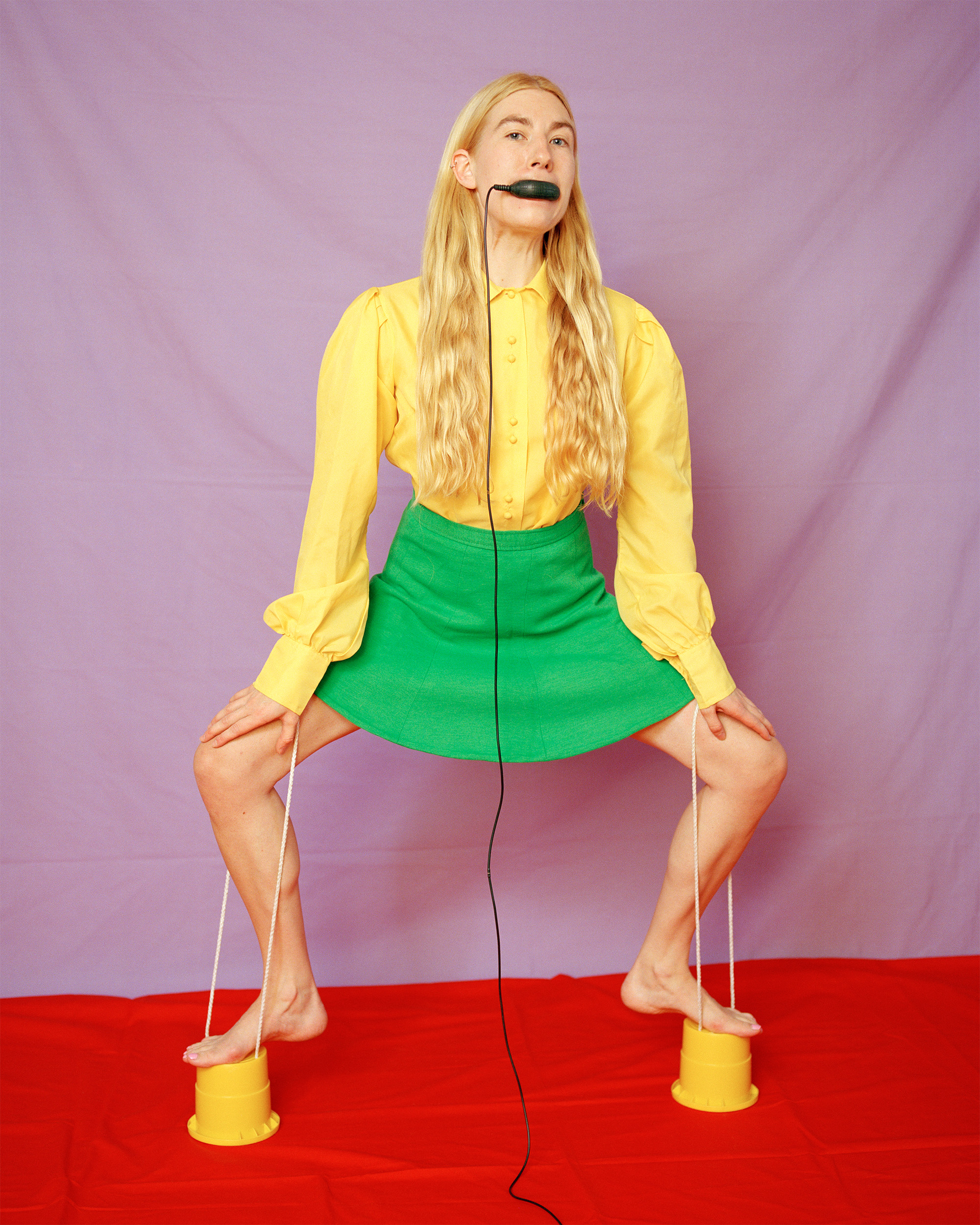 A woman with long blond hair stands on top of two plastic cups as stilts. The strings of the stilts are looped over her knees as she crouches slightly. She is wearing a bright yellow blouse and a green mini skirts. She holds the remote shutter cable in her mouth and looks straight at the camera. 