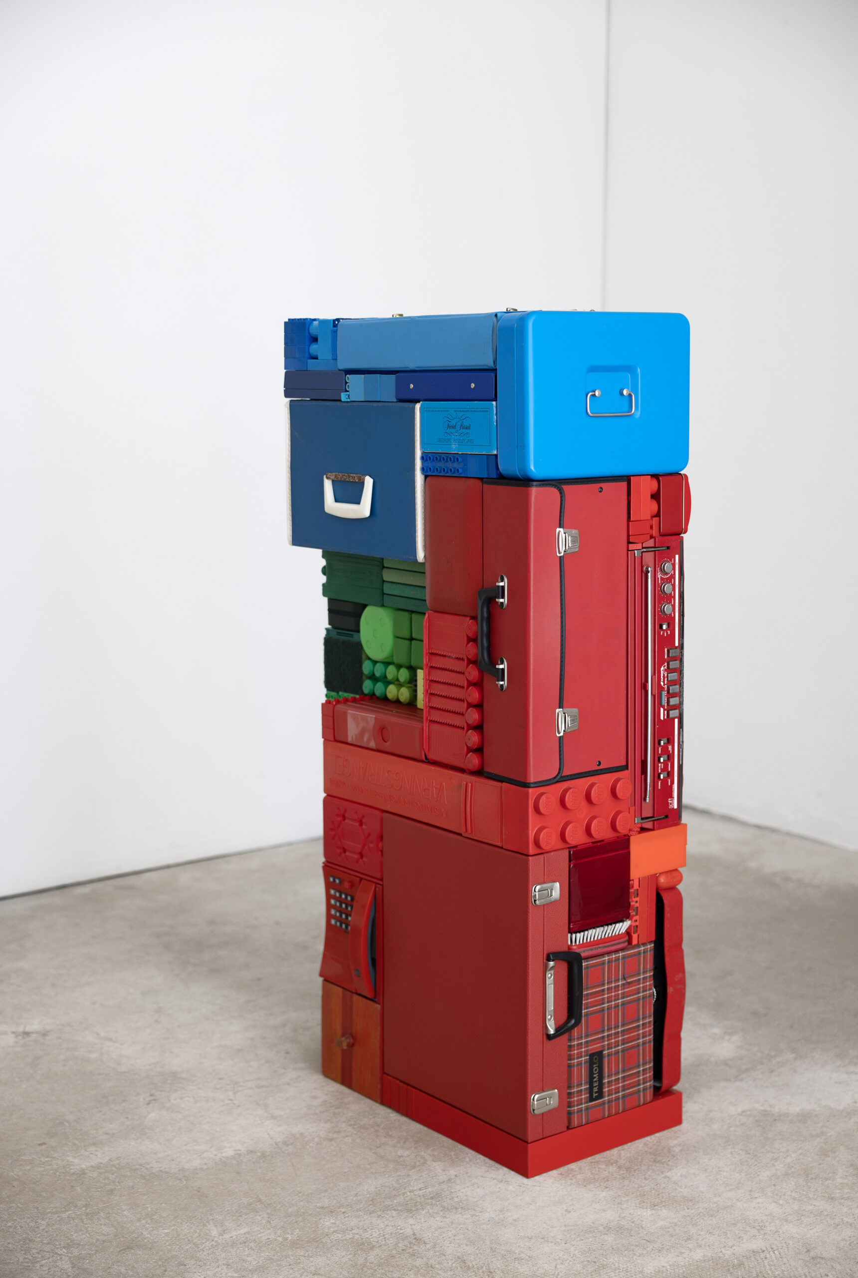 Filing boxes, storage boxes and giant pieces of lego are stacked on top of another tightly forming a neat stack. The top of the pile is blue, with green in the middle left and the majority of the stack is composed of red items. The stack stands in an empty, gallery space with white walls and cement flooring.