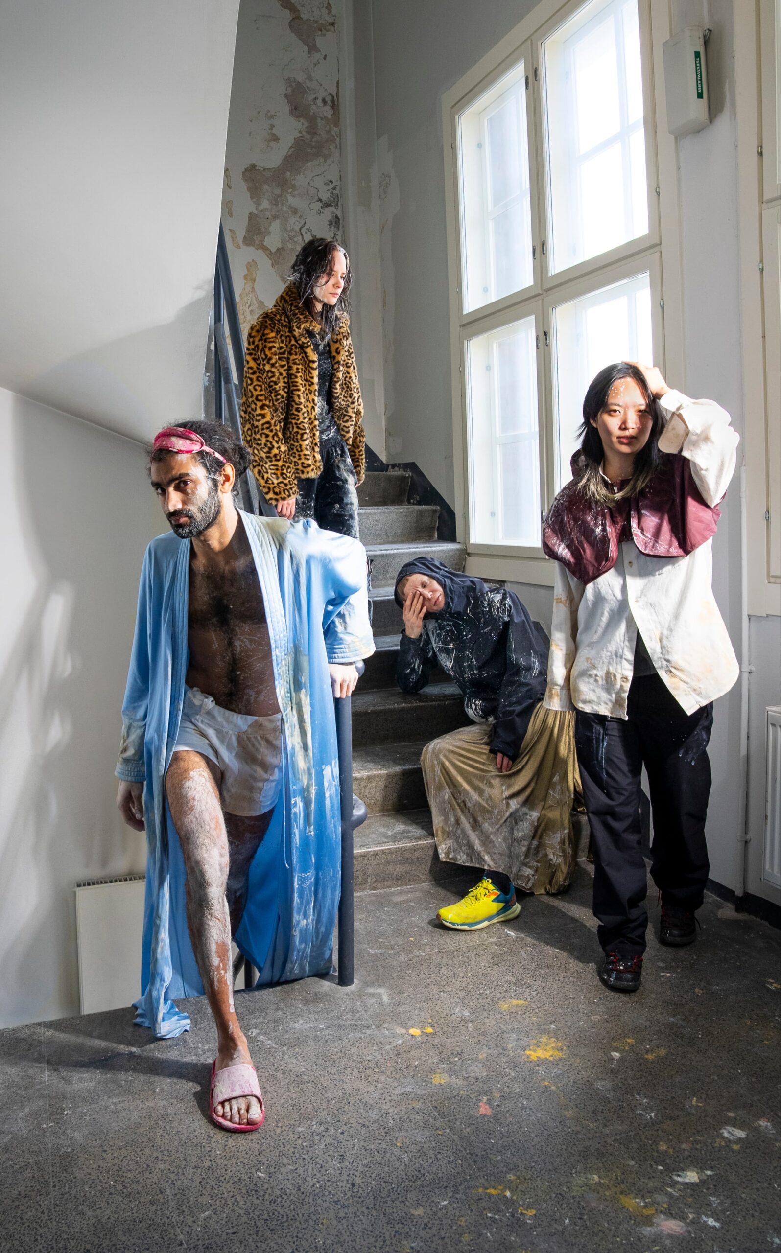 Four people stand in a window lit stairwell. thEY Some are wearing coats while the man to the forefront is wearing boxer shorts, an open dressing gown and an eye mask on his head. They all appear to have paint, coloured chalk or spices on them. There is also patches spattered across of it on the floor. Some of them have their hands to their heads or eyes as in distress or tiredness.