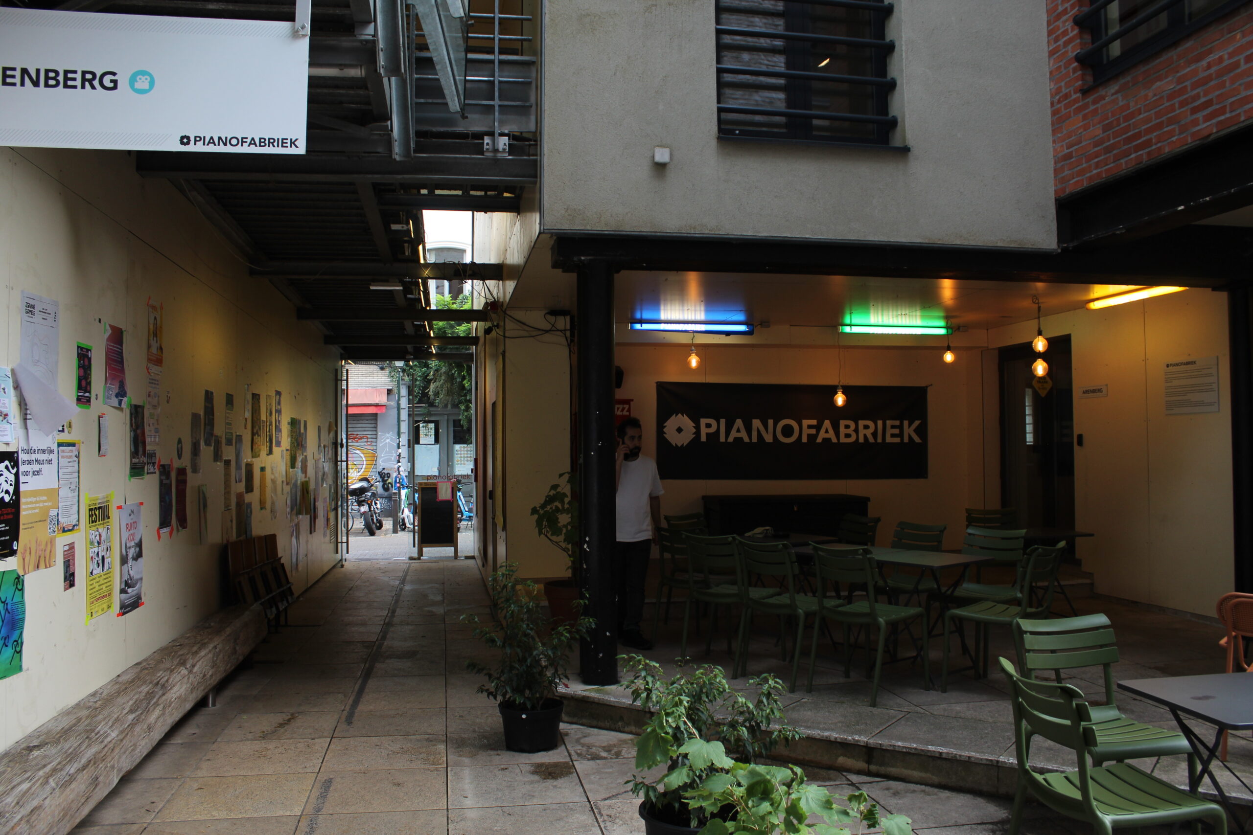 Pianofabriek. Photo by Oscar Arminen. The inner courtyard of Pianofabriek, posters line the wall to the left. Potted plants tables and chairs are places around the space. A sign reads Pianofabriek.