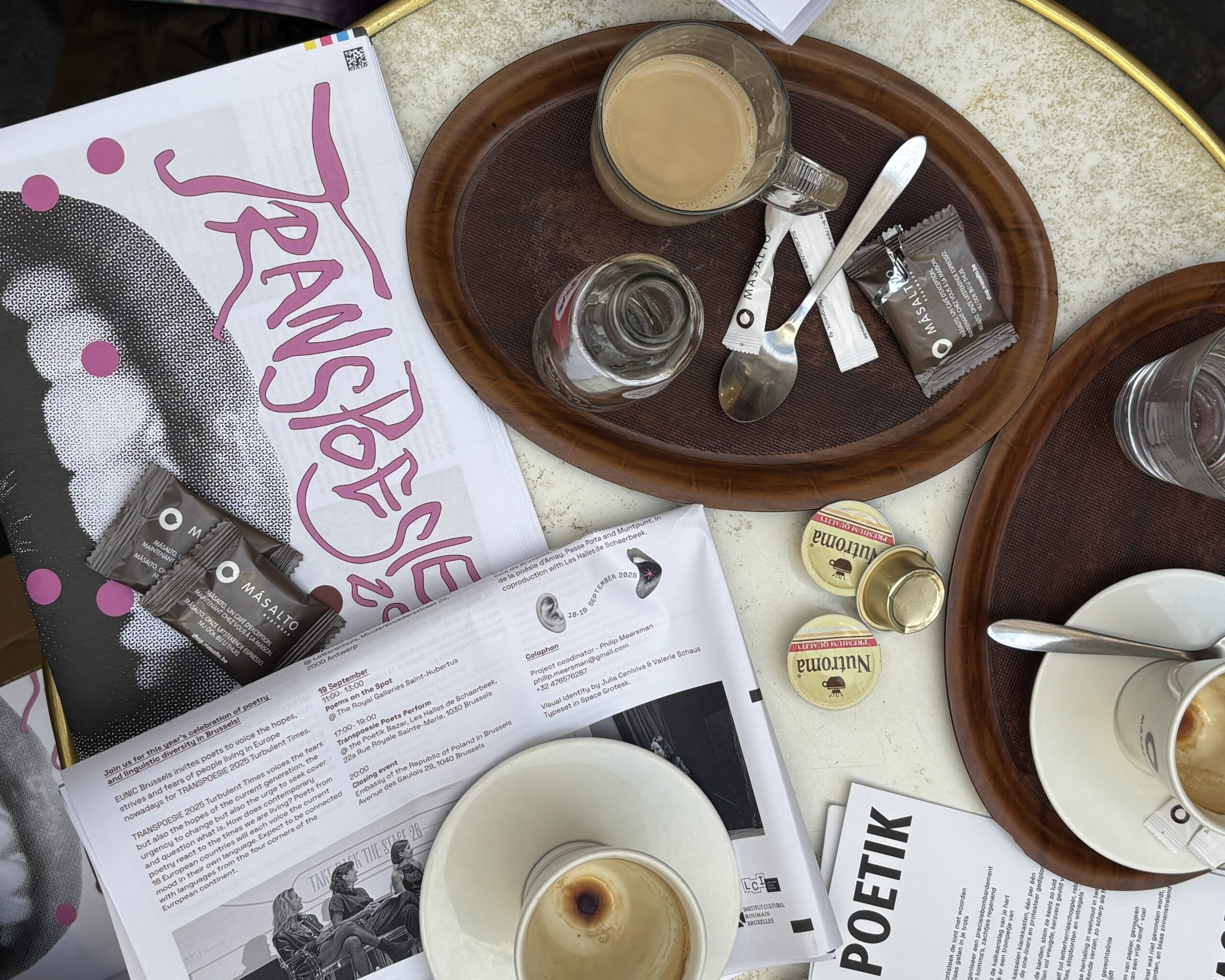 Transpoesie 2025. Photo Coffee cups, cutlery and newspapers cover a cafe table. Transpoesie is the title of one of the papers.