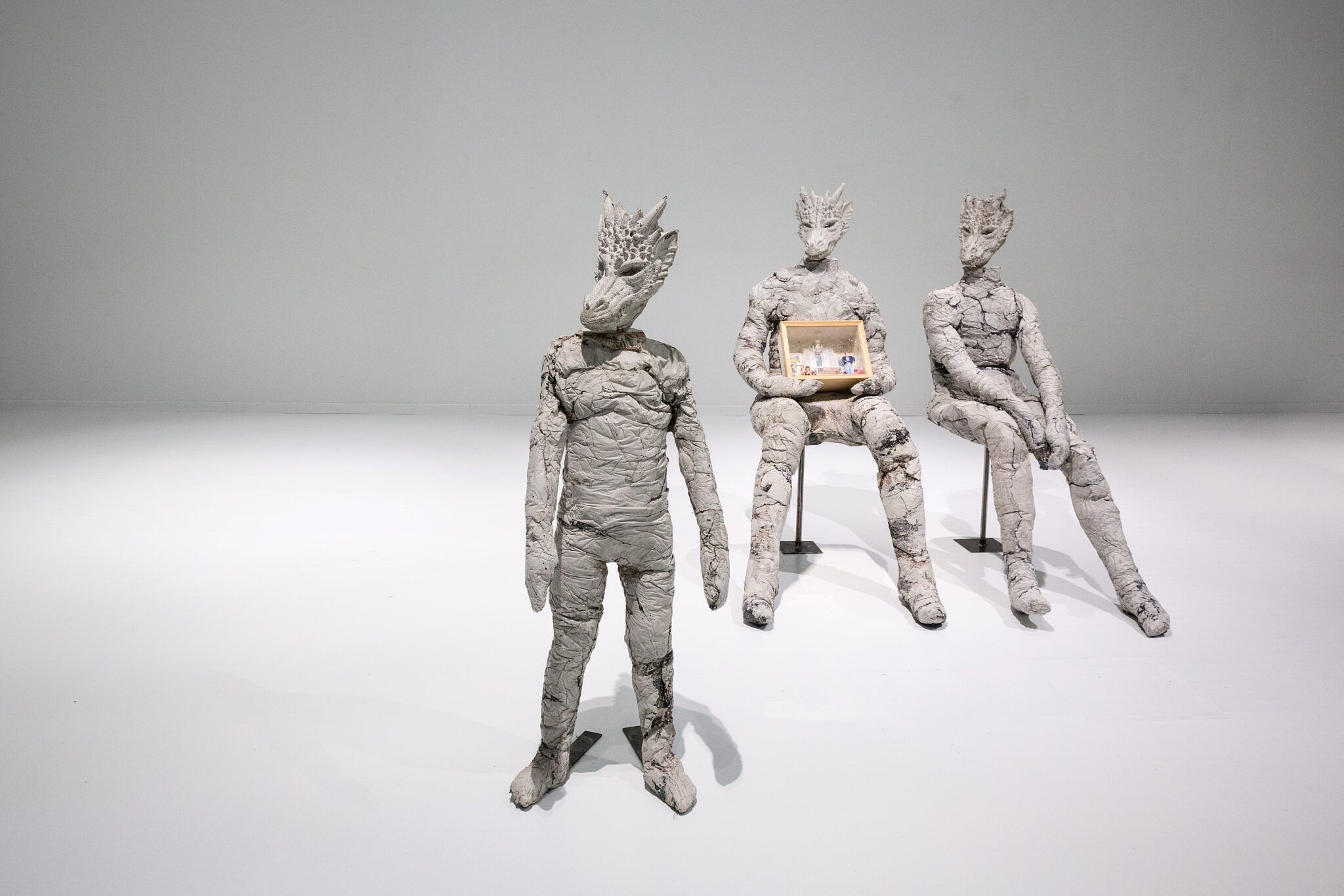Three grey sculpted humanoid figures with reptilian or dinosaur-like heads displayed in a white gallery space. One figure stands in the foreground, while two sit on a metal stand in the background — one of them holding a small framed display case resembling a dollhouse room. The sculptures have a rough, textured surface resembling clay or plaster.