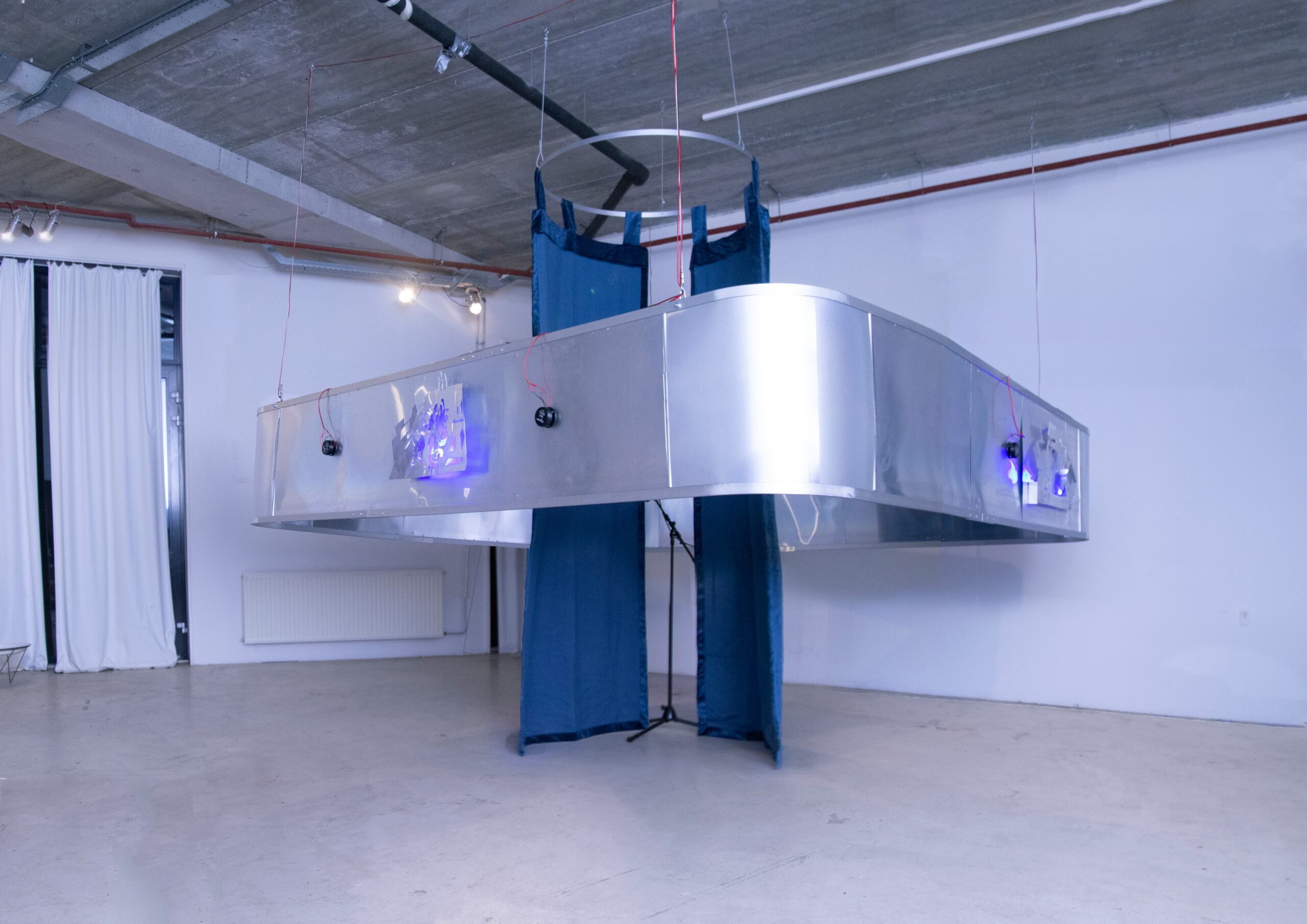 A suspended gallery installation featuring a polished aluminium rectangular frame with blue fabric panels, small speakers, and blue LED lights, hanging from a concrete ceiling by wires.