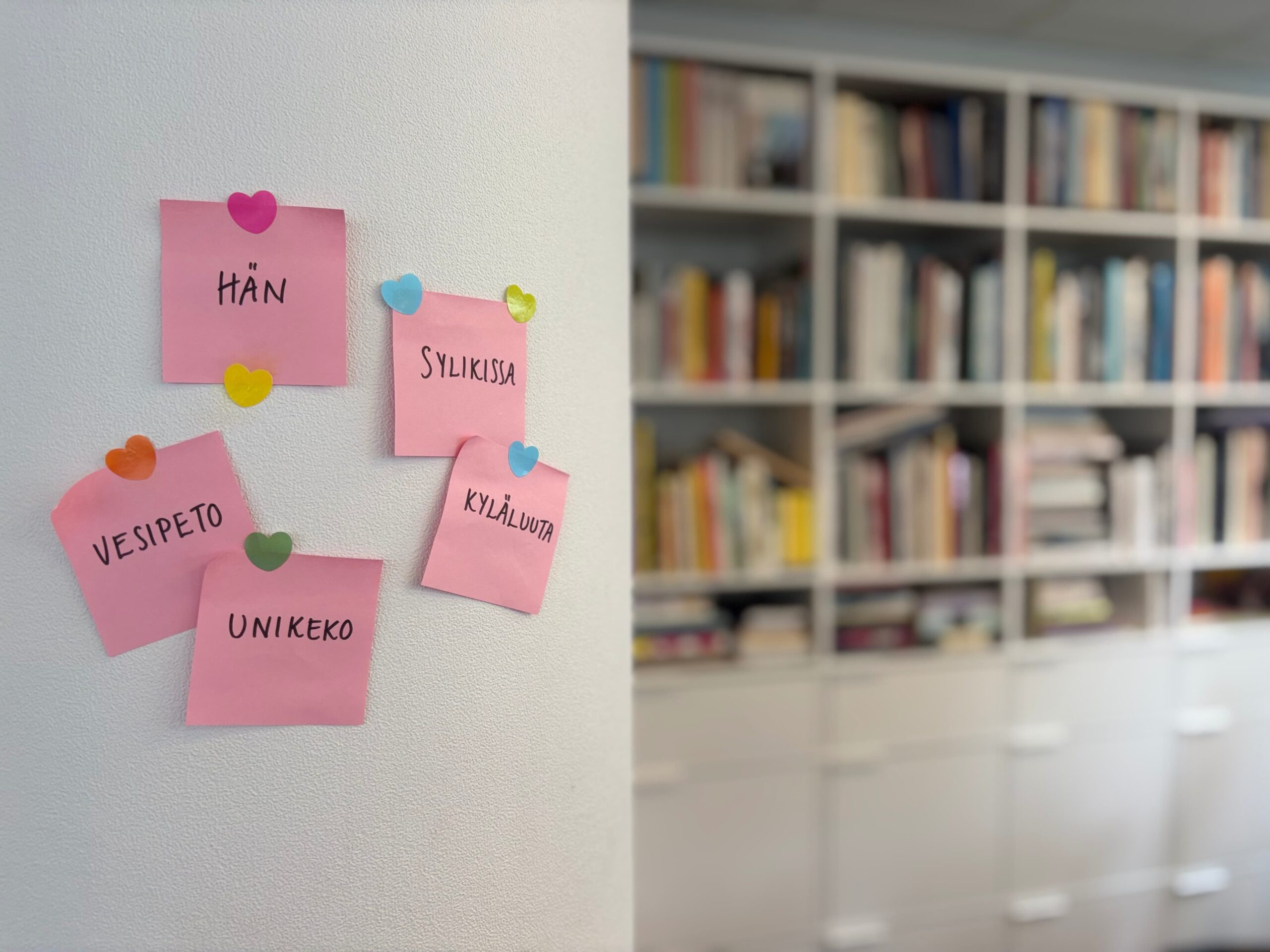 Words in Finnish are written on pink post-it's stuck on a white wall. In the background to the right there is a bookshelf out of focus.