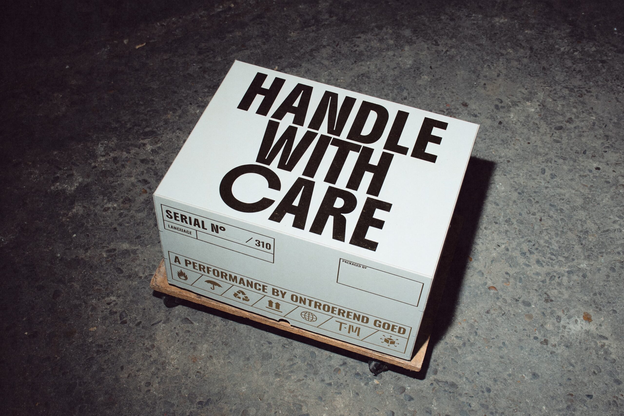 HTT – Ontroerend Goed – Handle with Care – Photo_ Joëlle Desmet-2 A white box sits on a wooden platform on wheels on a cement floor. "Handle with care" is written in all caps on the top of the box.