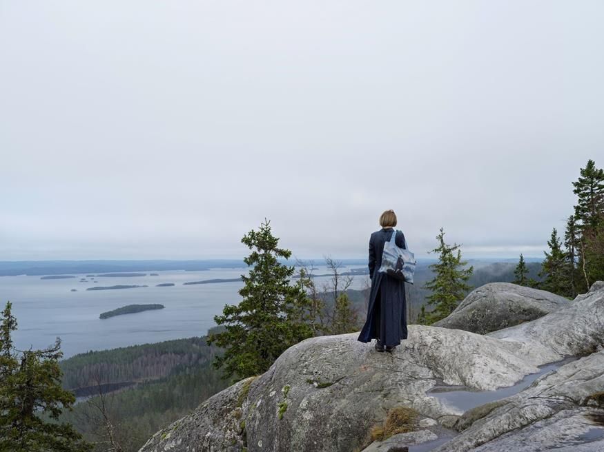 A woman stands on a rocky landscape looking out to sea in Finland. She wears a long navy flowing dress or jacket and carries a large bag on her shoulder. The image on the bag is of someone standing on rocks looking out at the horizon.