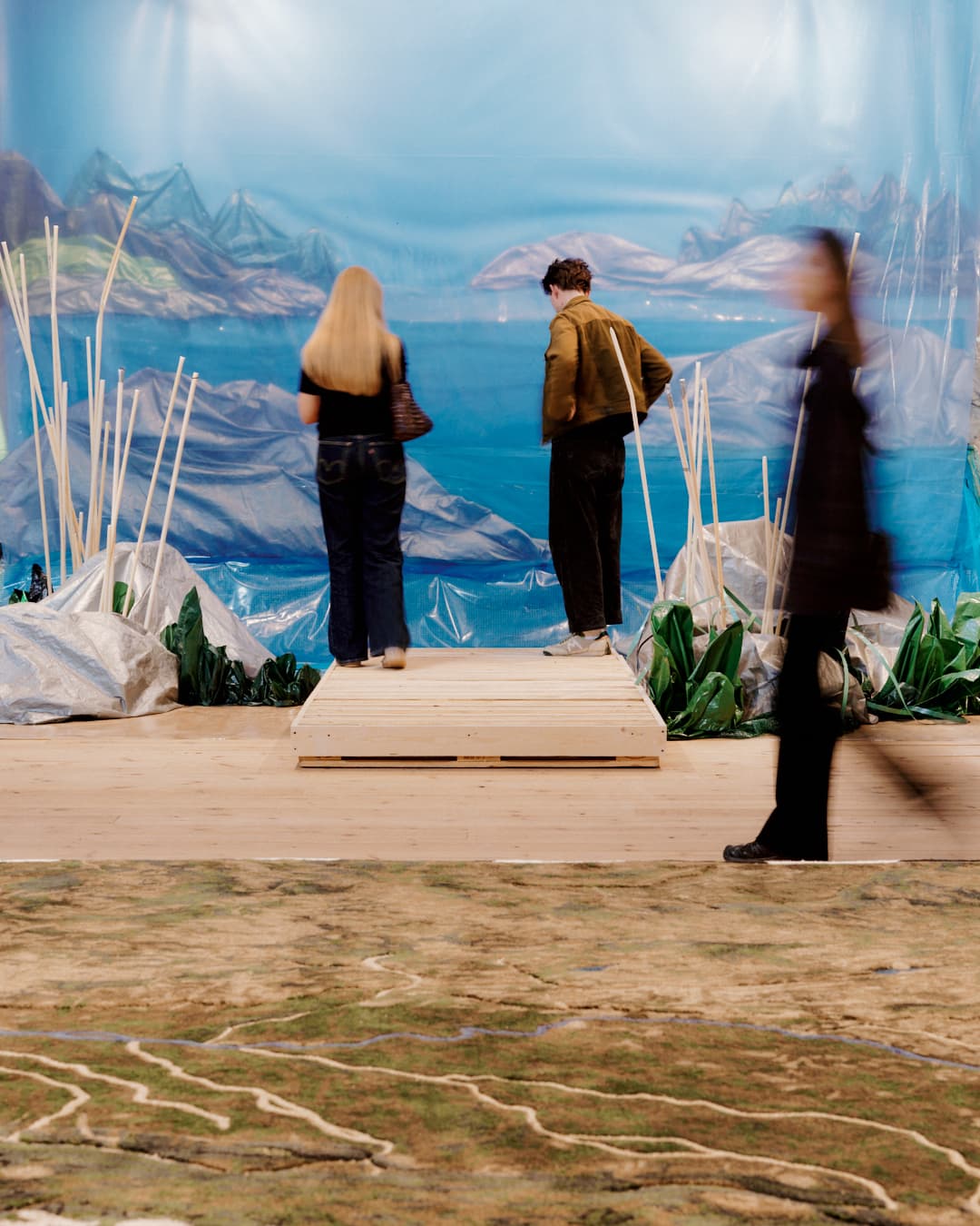 Two people are seen standing on a wooden platform looking at a lake or sea view with islands in the background. In the foreground a woman is walking but is blurred due to her movement. She walks behind a grassy, sandy, textured map-like topography on the floor.