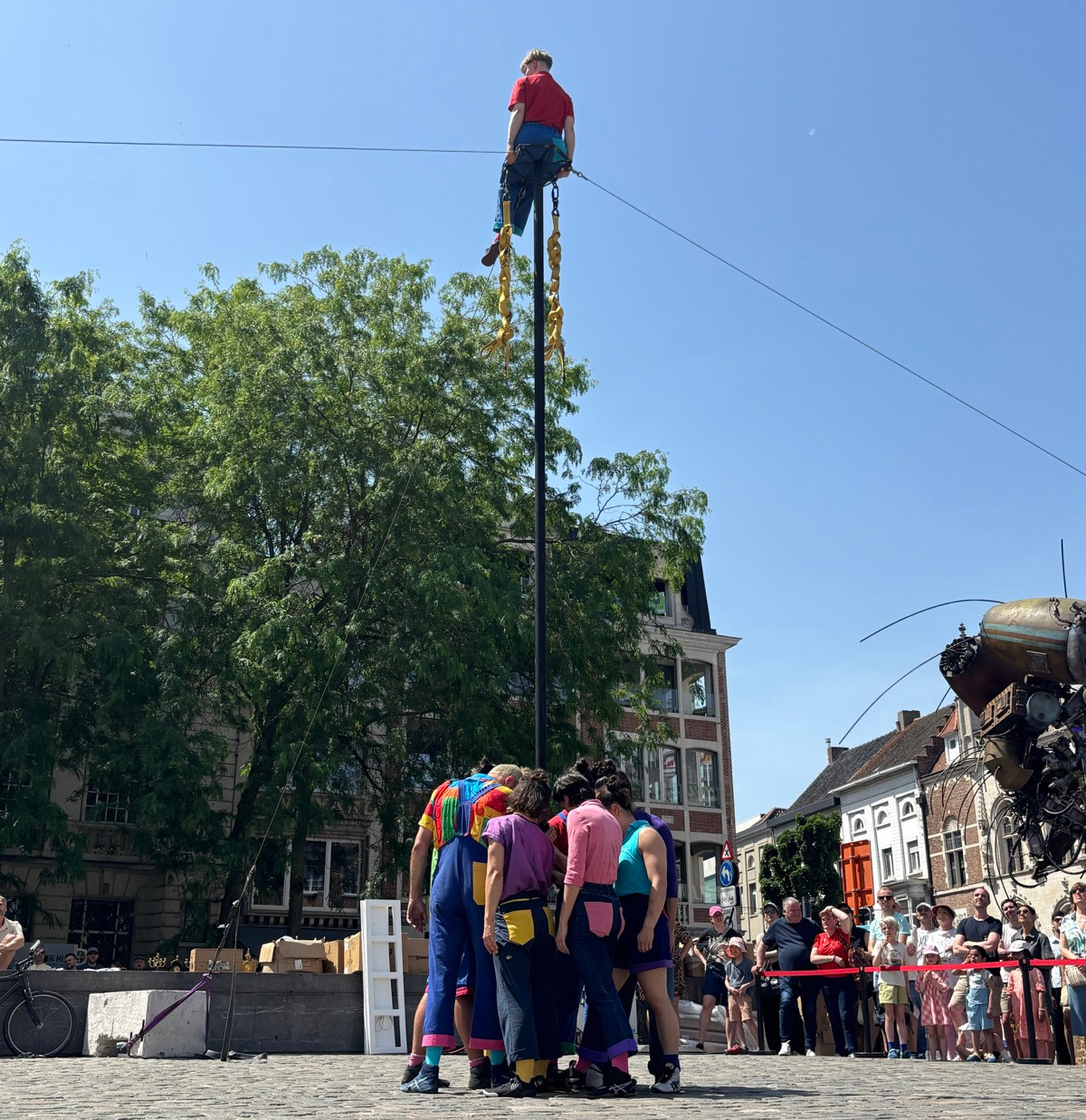 A group of circus artists stand in a circle in the street, above them a person balances on a high pole.