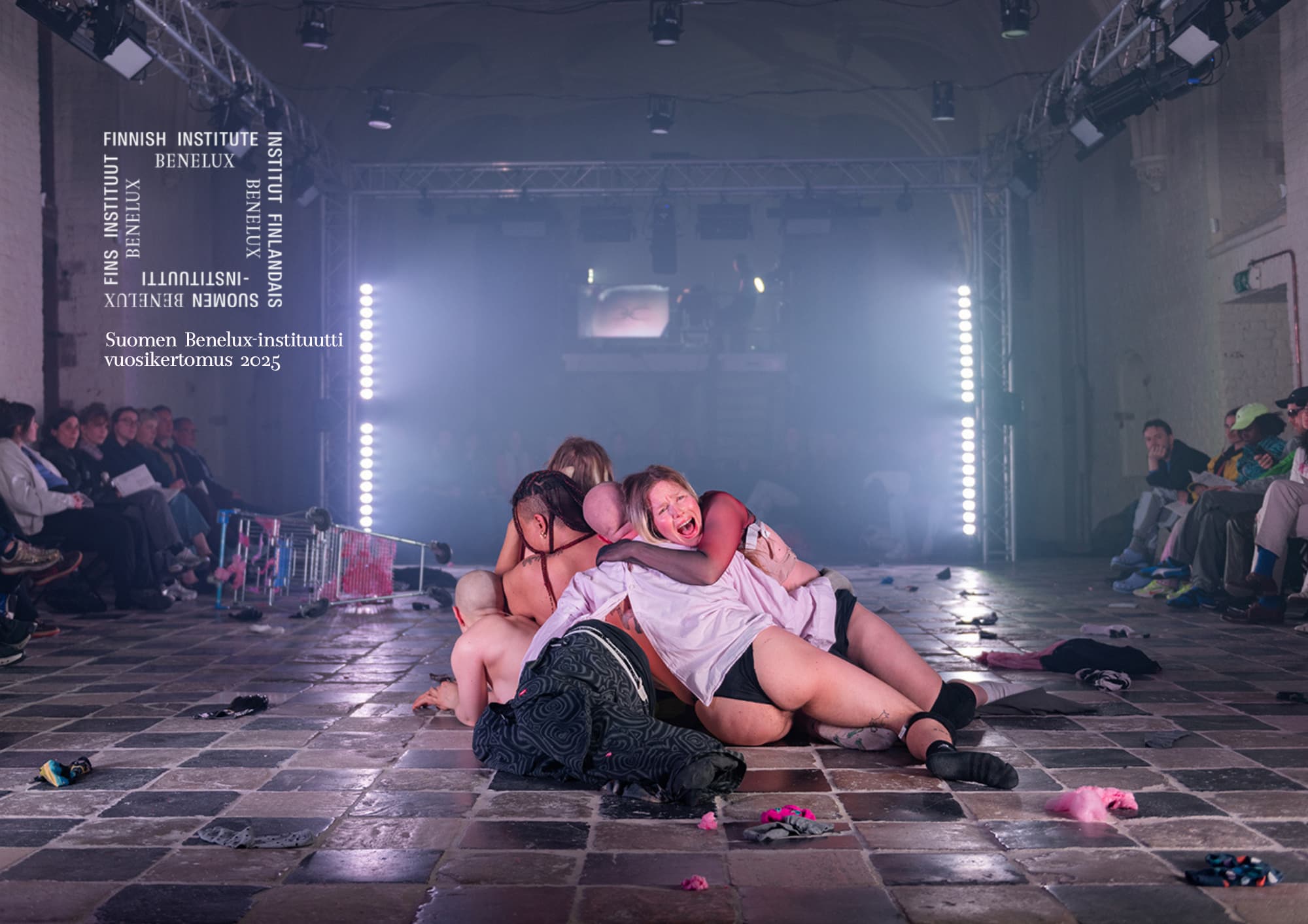 A group of performers lie, stretch and curl into each other on a black and white tiled floor. White strip lights line the two sides of the back wall. The audience is sits alone the two sides of the room facing each other across the performers.