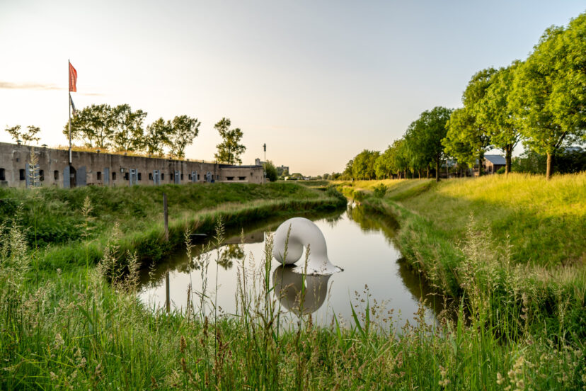 A large sculpture of a horn can be seen in a moat surrounding an old barrack. The nature is calm and serene.