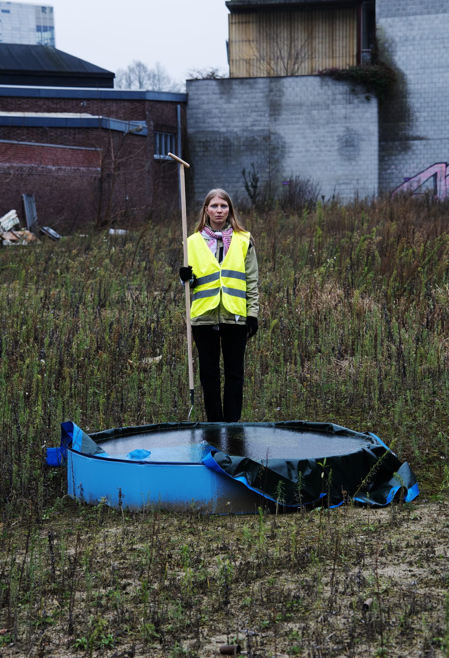 Vilma Vanhanen photo by Laetitia Delbachian A woman with long blind hair stands in an urban scrub wasteland behind a filled children's paddling pool. She is wearing a high visibility vest and is holding a long handled gardening tool.