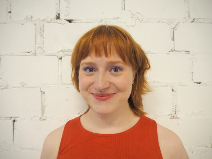 A woman with short ginger hair smiles at the camera. She is wearing a sleeveless orange top. She is standing against a brick wall painted white.
